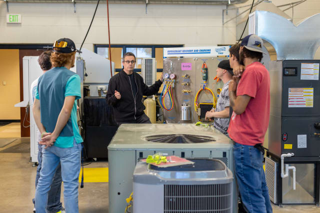 This image depicts five students and one teacher around an air conditioning unit learning how to test and repair. until
