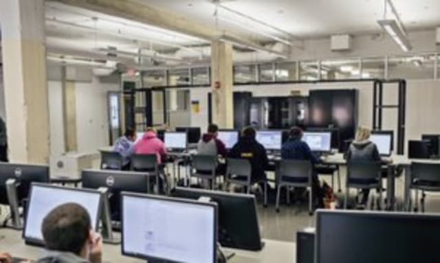 This image depicts a computer lab with computers and students sitting at computers working.