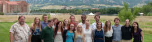 A large group of space science students stand on East Campus of CU Boulder with the majestic Flatirons behind them. 