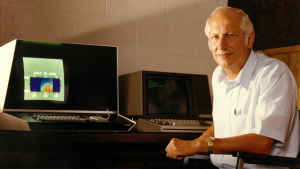 An old historic photo of a male scientist sitting in front of a desk with large vintage computers. 