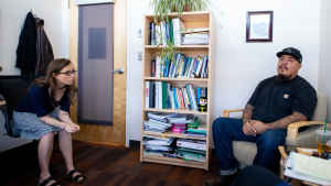 A female mental health clinican leans over in her chair to listen to a veteran sitting across from her during a therapy session. A bookcase filled with books and plants is against the wall. 