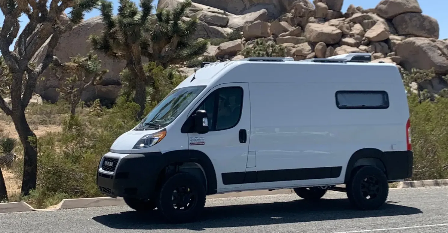 A sleek white van with black trim parked in a sparse desert environment featuring rock formations and Joshua trees under a clear blue sky.
