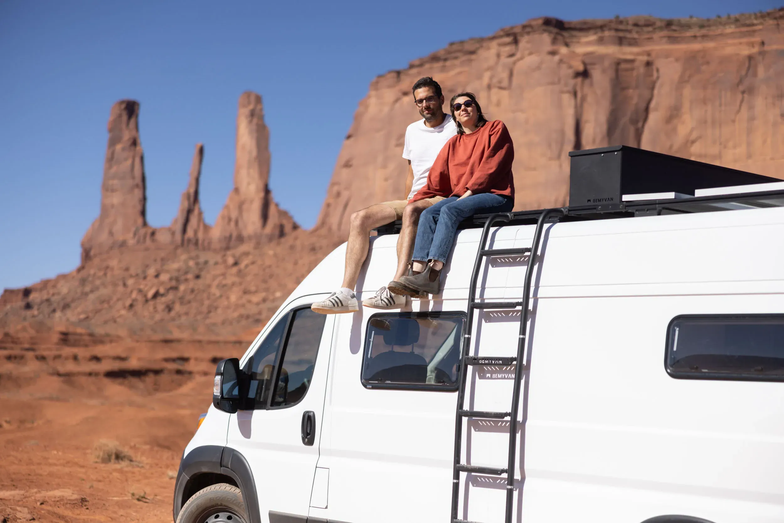 A couple sitting on the roof of a white camper van, smiling as they admire red rock formations in a desert landscape under a clear blue sky.
