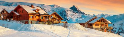 Wooden chalets in Alpe d'Huez during sunset, with snow-covered mountains in the background.