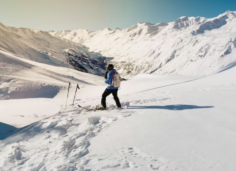 Skier standing at the edge of a snowy slope, overlooking a vast alpine valley with snow-covered peaks under a clear blue sky