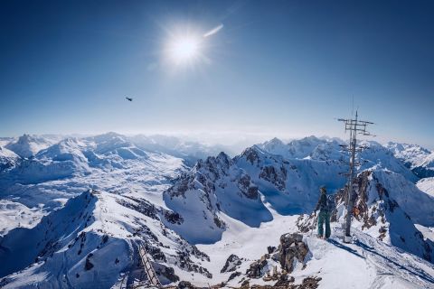 Snowy mountain peaks under a bright sun with a skier and bird in view.