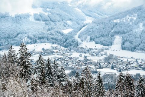 Snow-covered alpine village nestled between forested mountains, with ski runs visible.
