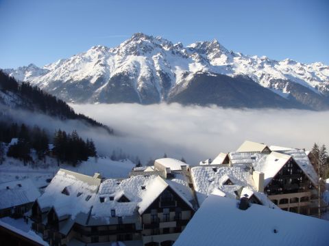 Snow-covered mountain village with chalet-style buildings in the foreground, surrounded by a misty valley and majestic, snow-capped mountain peaks in the background on a clear winter day in the French Alps