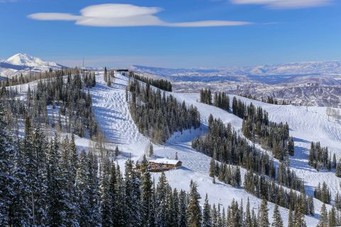 Snow-covered slopes lined with pine trees in Aspen, Colorado, with a ski lodge nestled at the base and panoramic views of the Rocky Mountains under a clear blue sky.