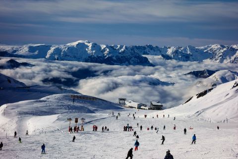 Skiers descend a snowy slope in a French Alps resort with a dramatic cloud inversion below and rugged peaks rising in the distance.