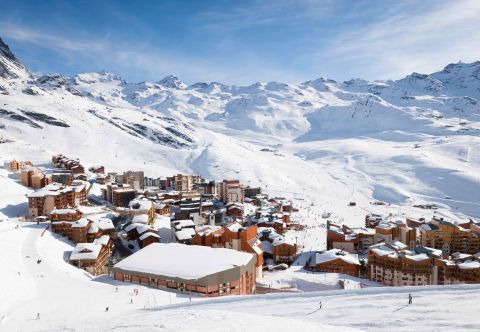 Panoramic view of Val Thorens resort nestled high in the French Alps, with snow-covered buildings and skiers heading straight onto expansive alpine slopes under a bright blue sky