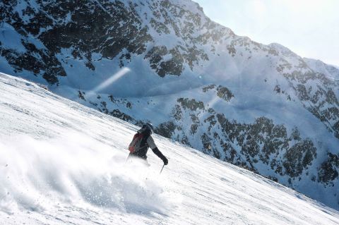 Skier carving through fresh powder on a steep, sunlit slope, surrounded by dramatic alpine peaks and rugged mountain terrain.