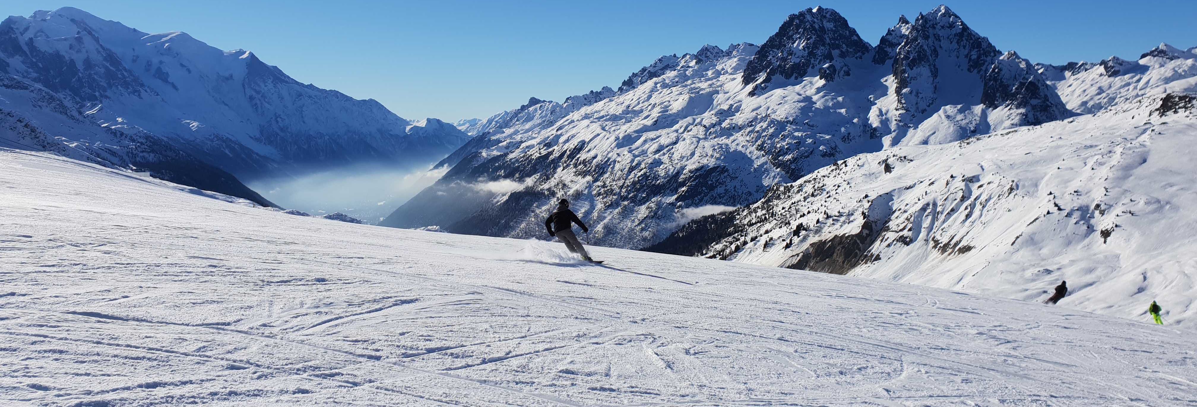 A skier on a wide, sunny piste in Chamonix with the Mont Blanc massif behind and cloud filling the valley