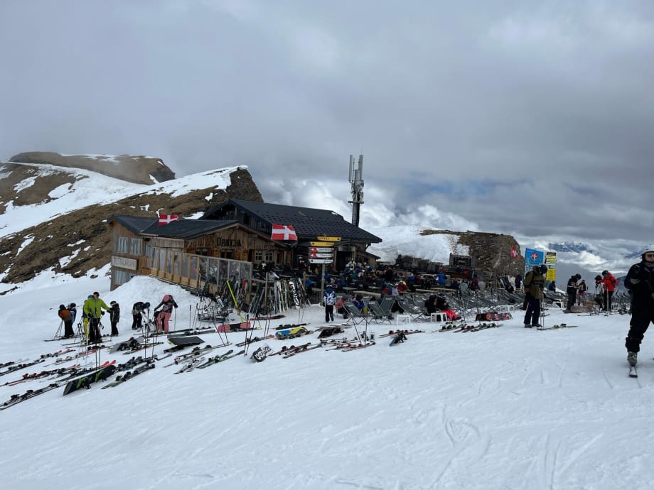 Skiers and snowboarders gathered outside a wooden mountain restaurant at the Swiss border, with skis propped up in the snow