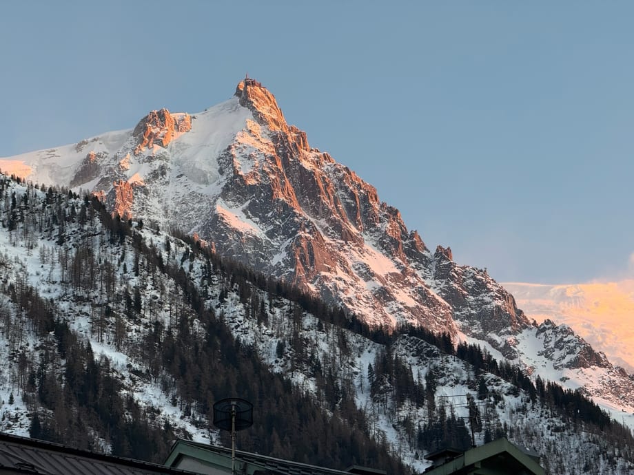 The rocky spires of Les Drus lit orange at sunset above forested slopes and a chalet roof in Chamonix
