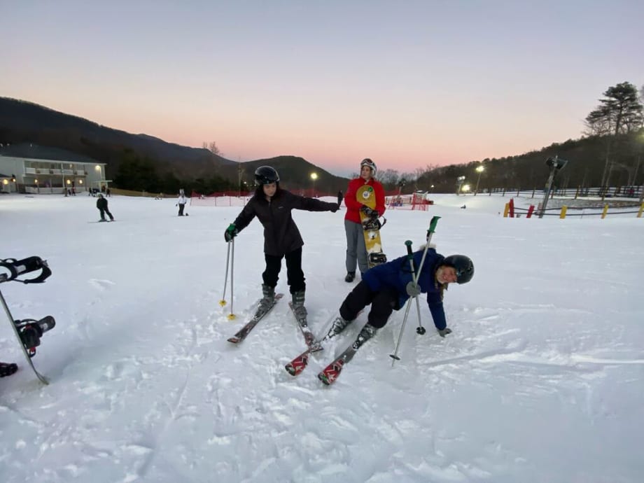 Skiers on a wide beginner slope at Snowshoe Mountain at sunset, one of them sitting down in the snow mid-fall, with the resort buildings behind