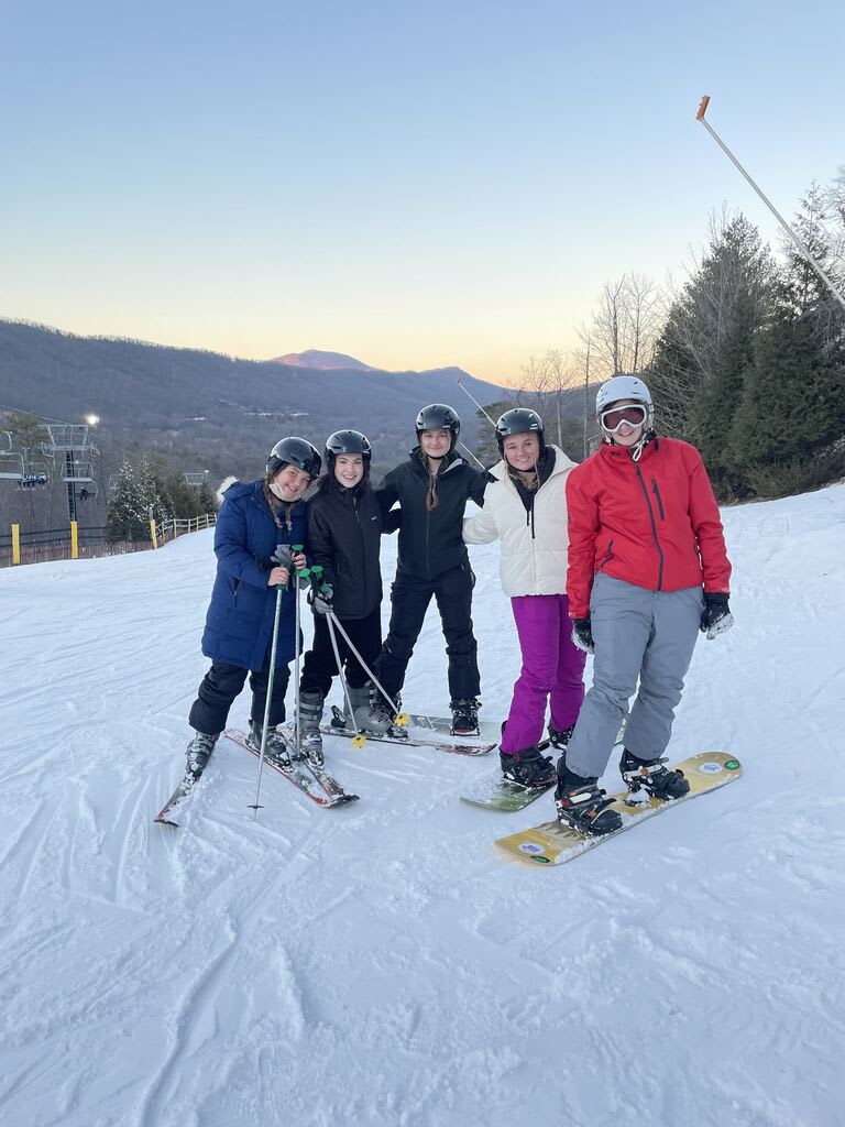 Five friends standing in a line on the snow at Snowshoe Mountain at dusk, with a wooded ridgeline and an orange sky behind them