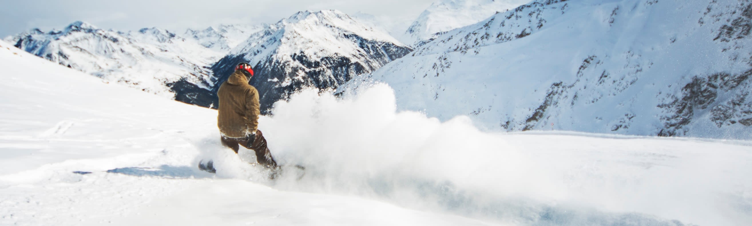 Snowboarder carving through fresh powder on a snowy mountain slope with alpine peaks in the background.