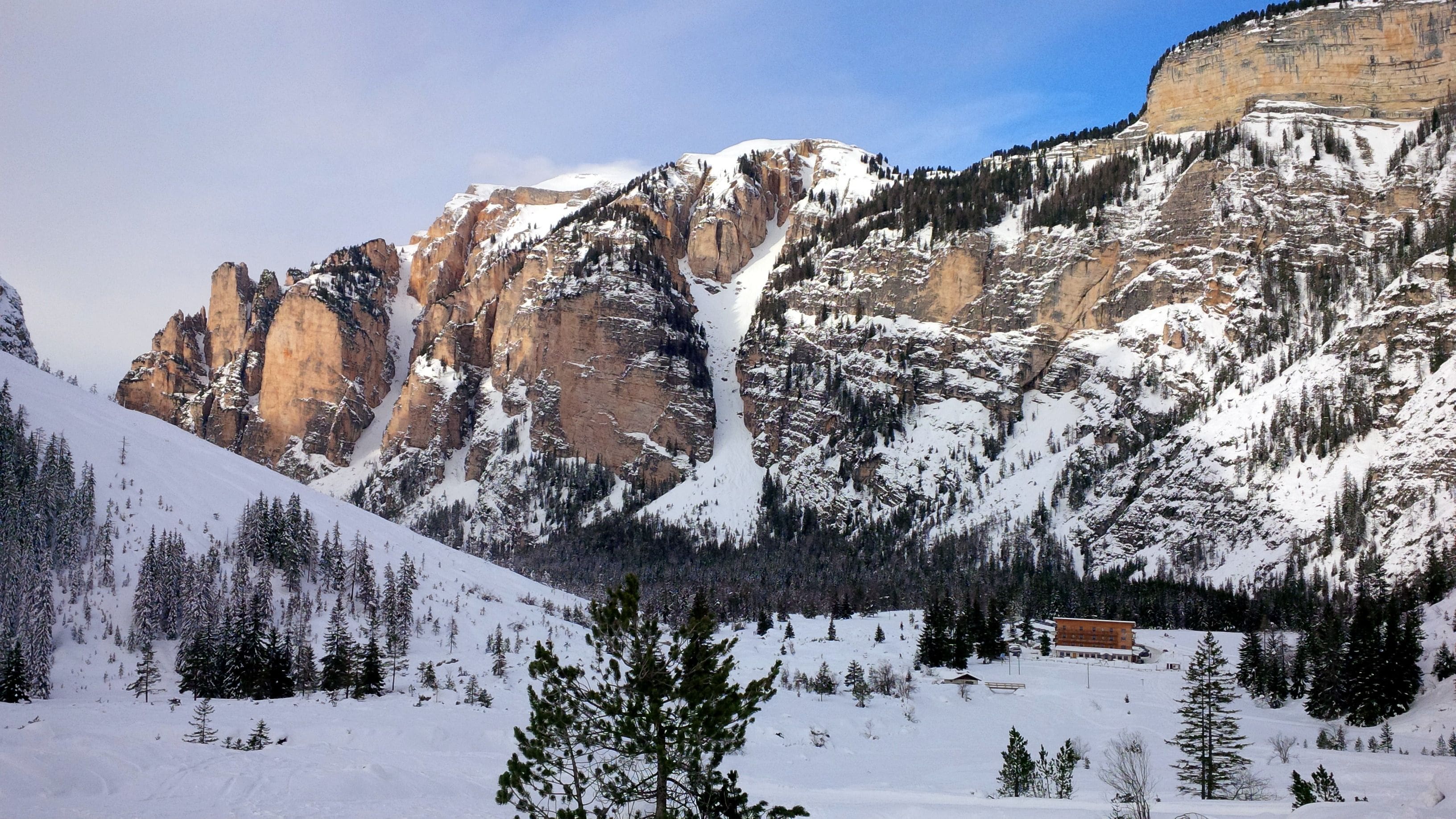 Majestic rocky cliffs with snow-covered pine trees and a snowy cabin in a valley.