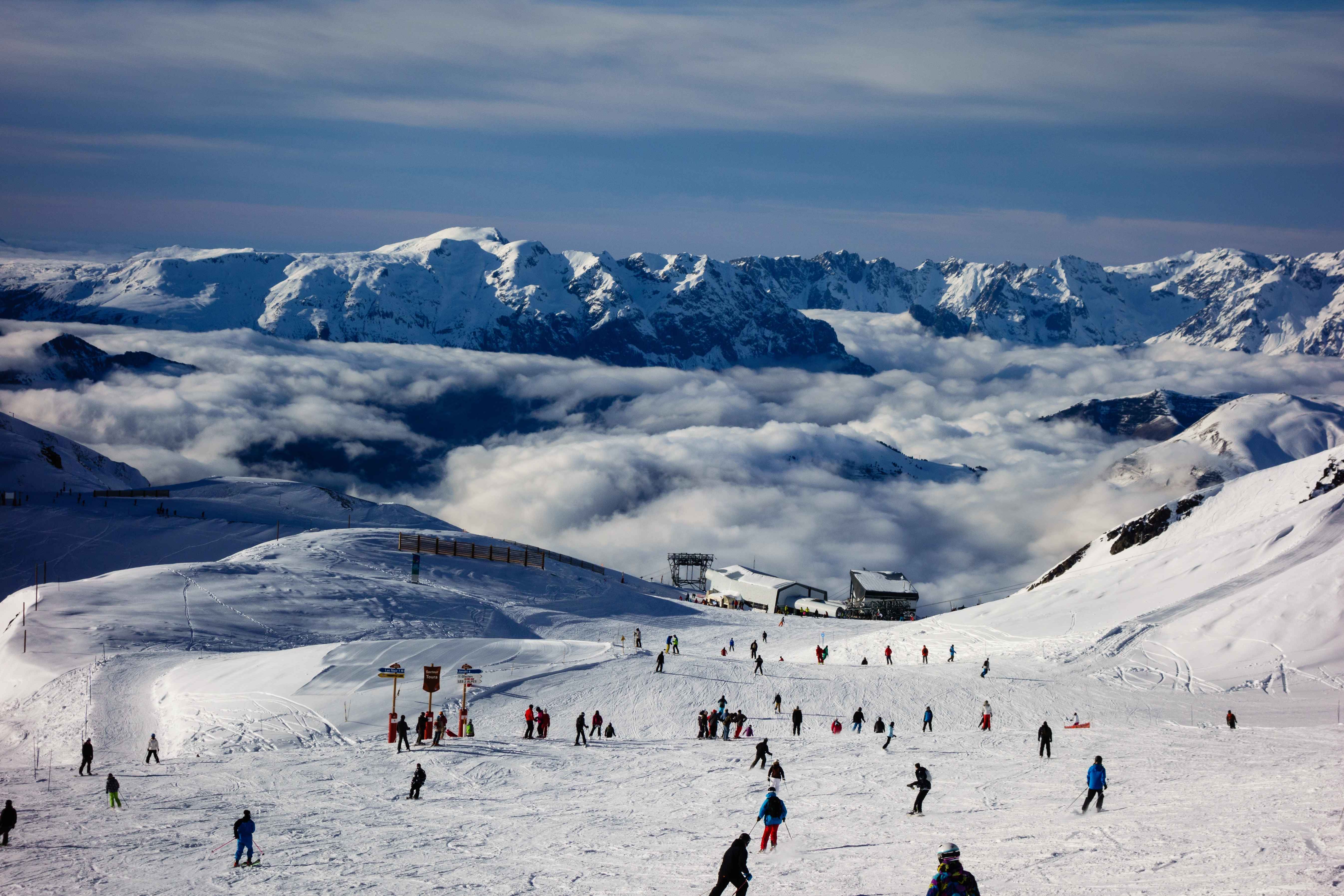 Skiers descend a snowy slope in a French Alps resort with a dramatic cloud inversion below and rugged peaks rising in the distance.