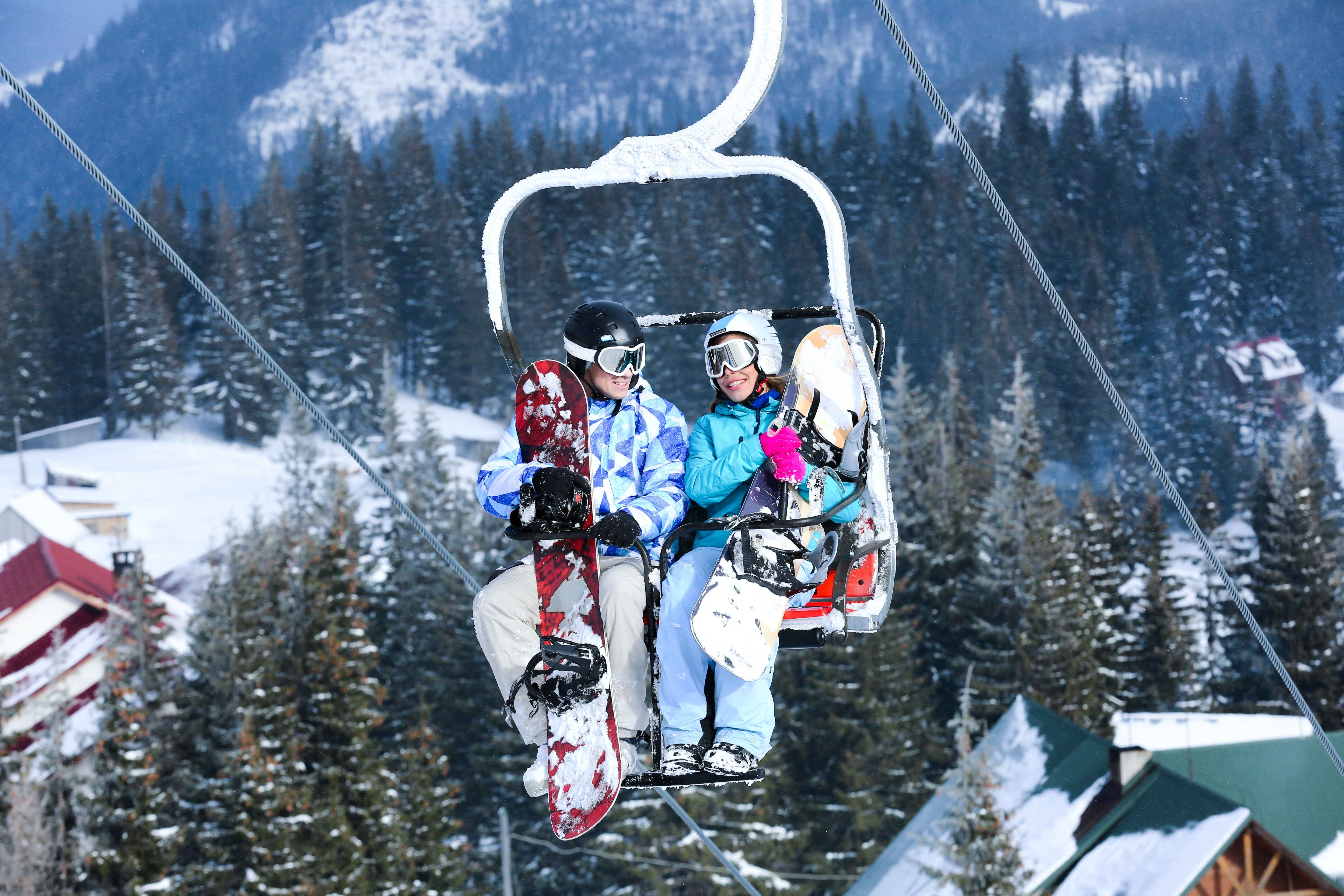Two snowboarders riding a snowy ski lift, smiling and chatting against a backdrop of pine trees and mountain lodges in a picturesque winter resort.