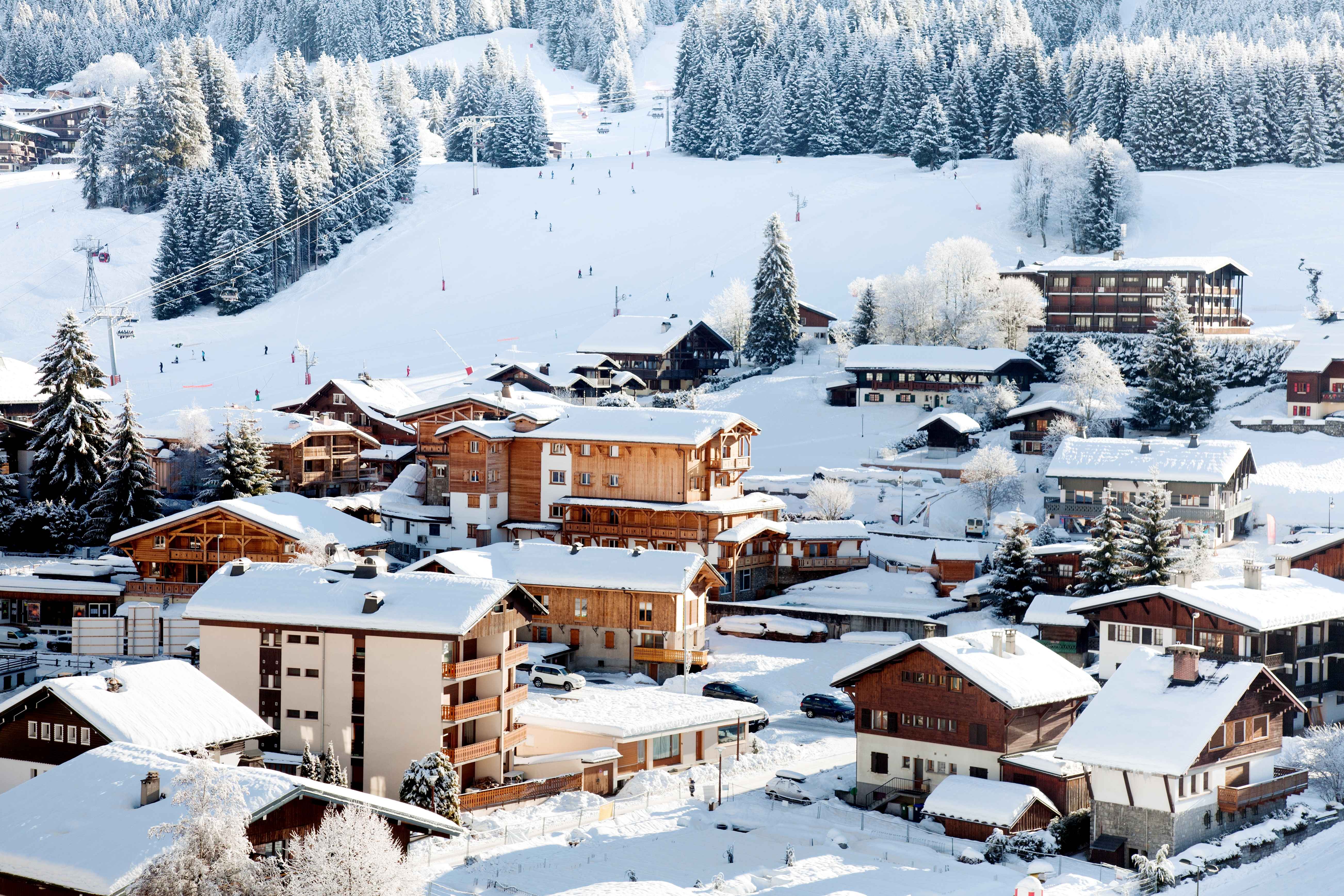Charming chalets blanketed in snow sit at the foot of tree-lined slopes, as skiers descend gently through the Les Gets ski resort beneath a bright winter sun.