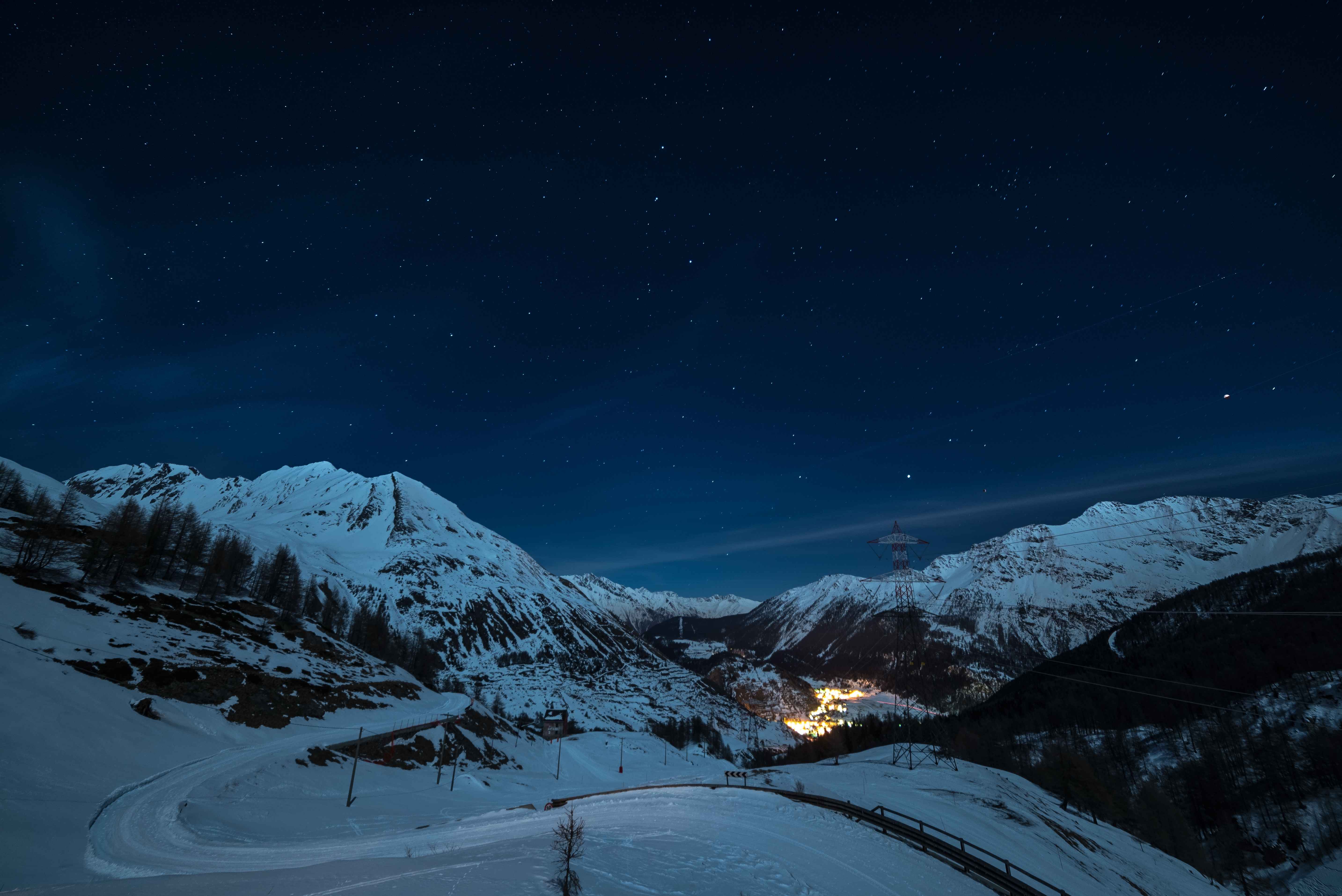A quiet winter road curves through the snowy Alps above La Thuile, where the village glows warmly in the valley below under a starlit sky offering a magical contrast between alpine serenity and cosy mountain life