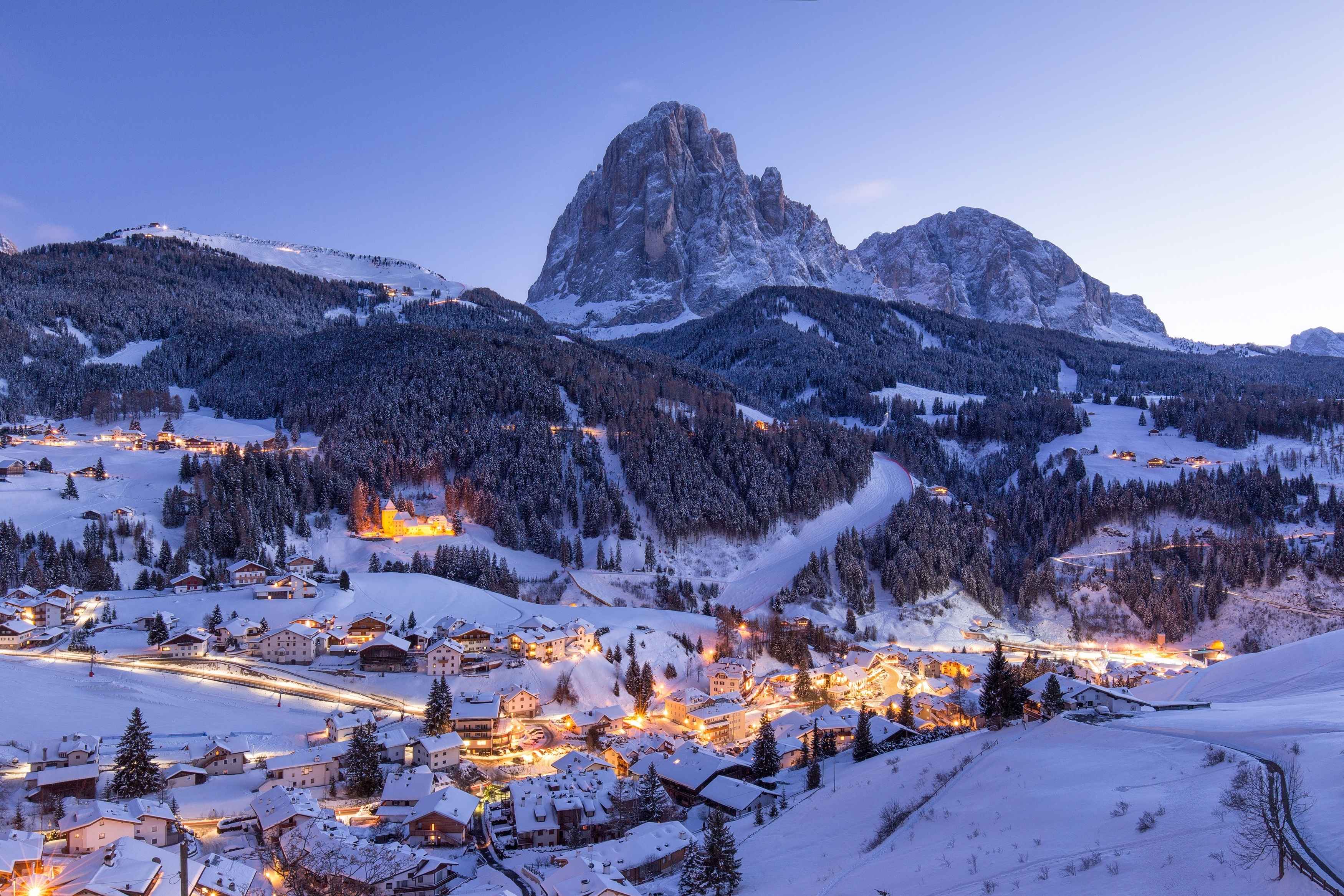A picturesque winter evening in the Dolomites, where a snow-covered village glows warmly under the towering peaks and forested slopes, creating a serene alpine scene