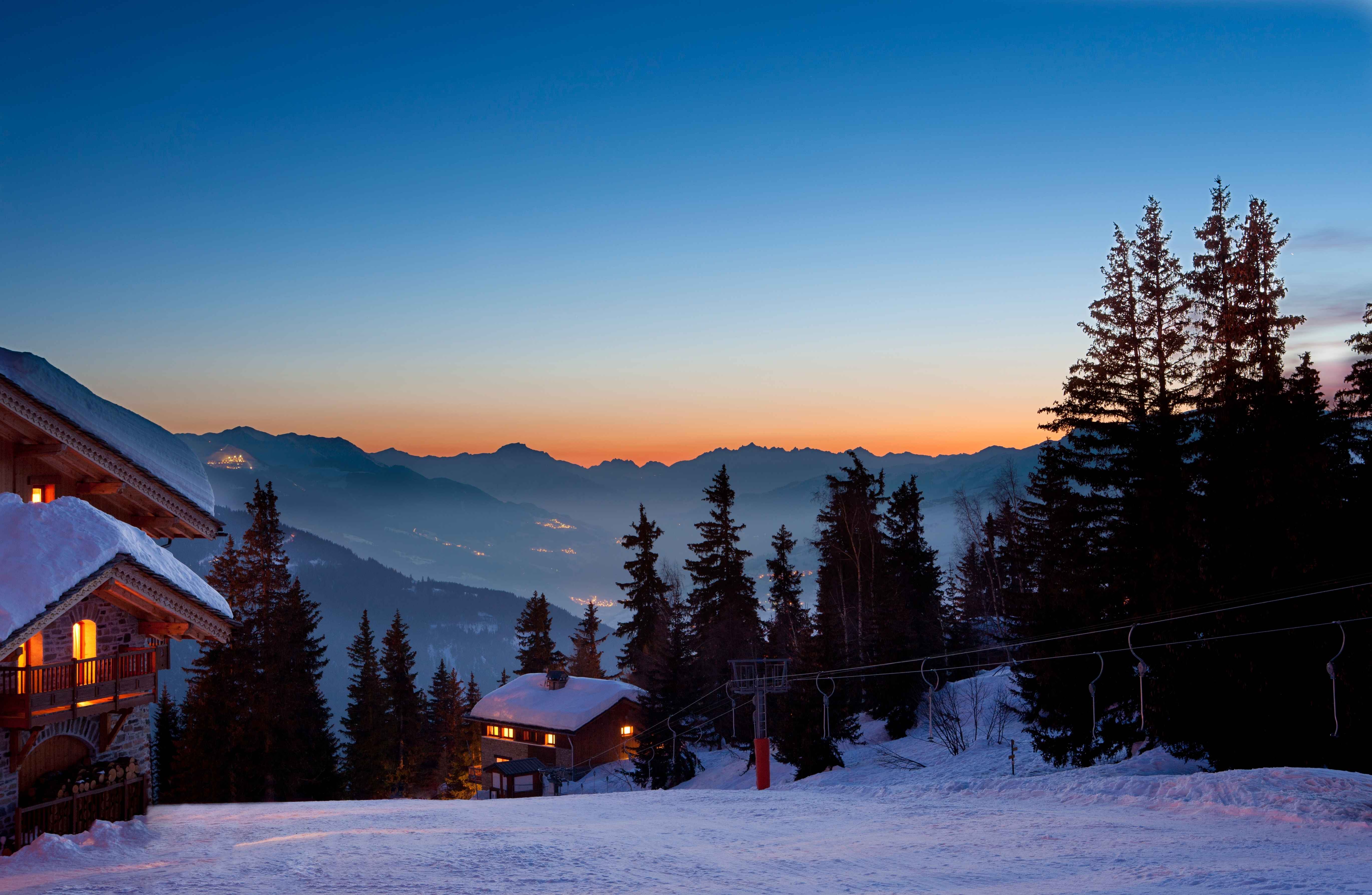 Snow-covered ski resort at dusk with glowing chalet lights, silhouetted pine trees, and mountain views under a twilight sky.