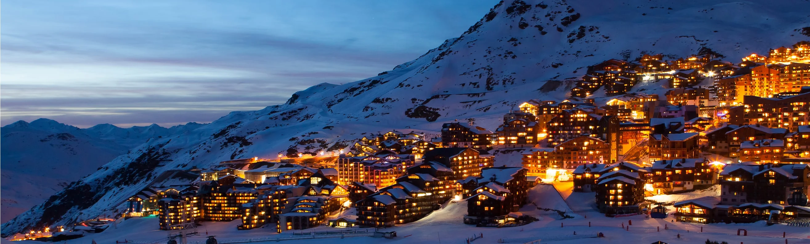 Val Thorens ski resort at dusk, with glowing lights on the mountainside and snow-covered chalets.