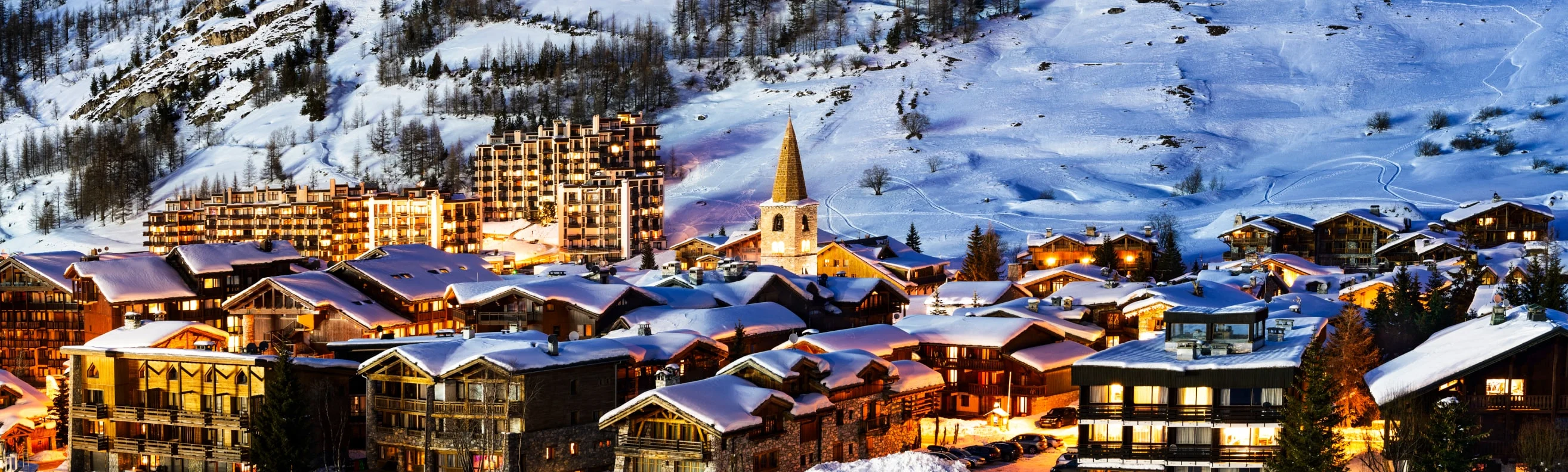 Val d'Isère nestled in the snow-covered mountains, illuminated at night, with traditional wooden chalets and modern lodgings creating a cozy, vibrant winter atmosphere