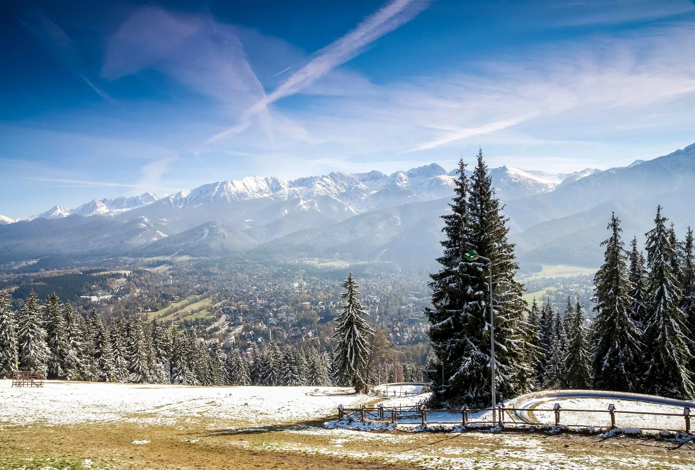Winter landscape of Zakopane, Poland, with snow-dusted pine trees, a picturesque valley, and the Tatra Mountains in the background under a clear blue sky.