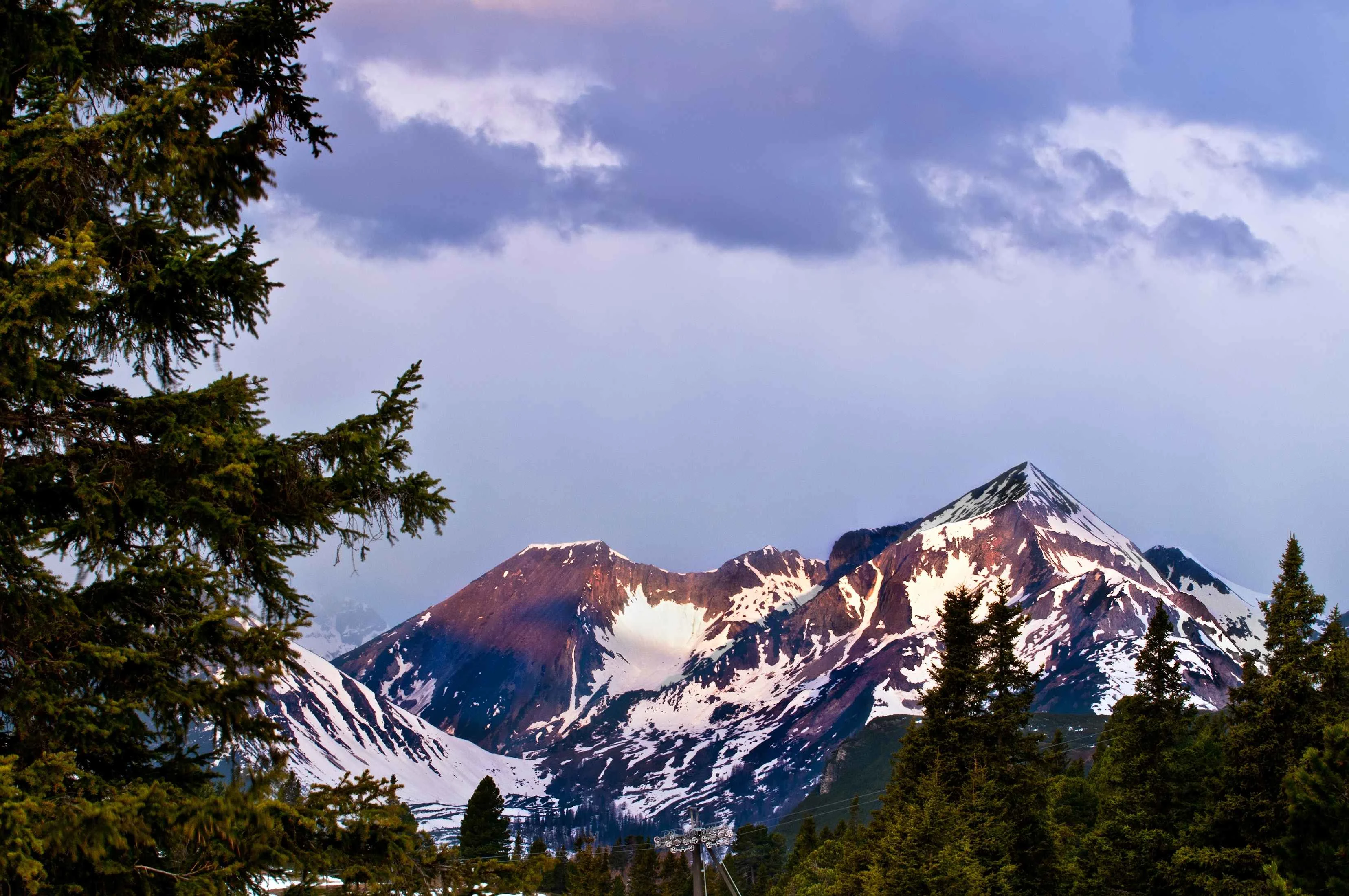 Snow-capped peaks of the Austrian Alps at dusk, with evergreen trees framing the rugged mountain landscape under a dramatic sky