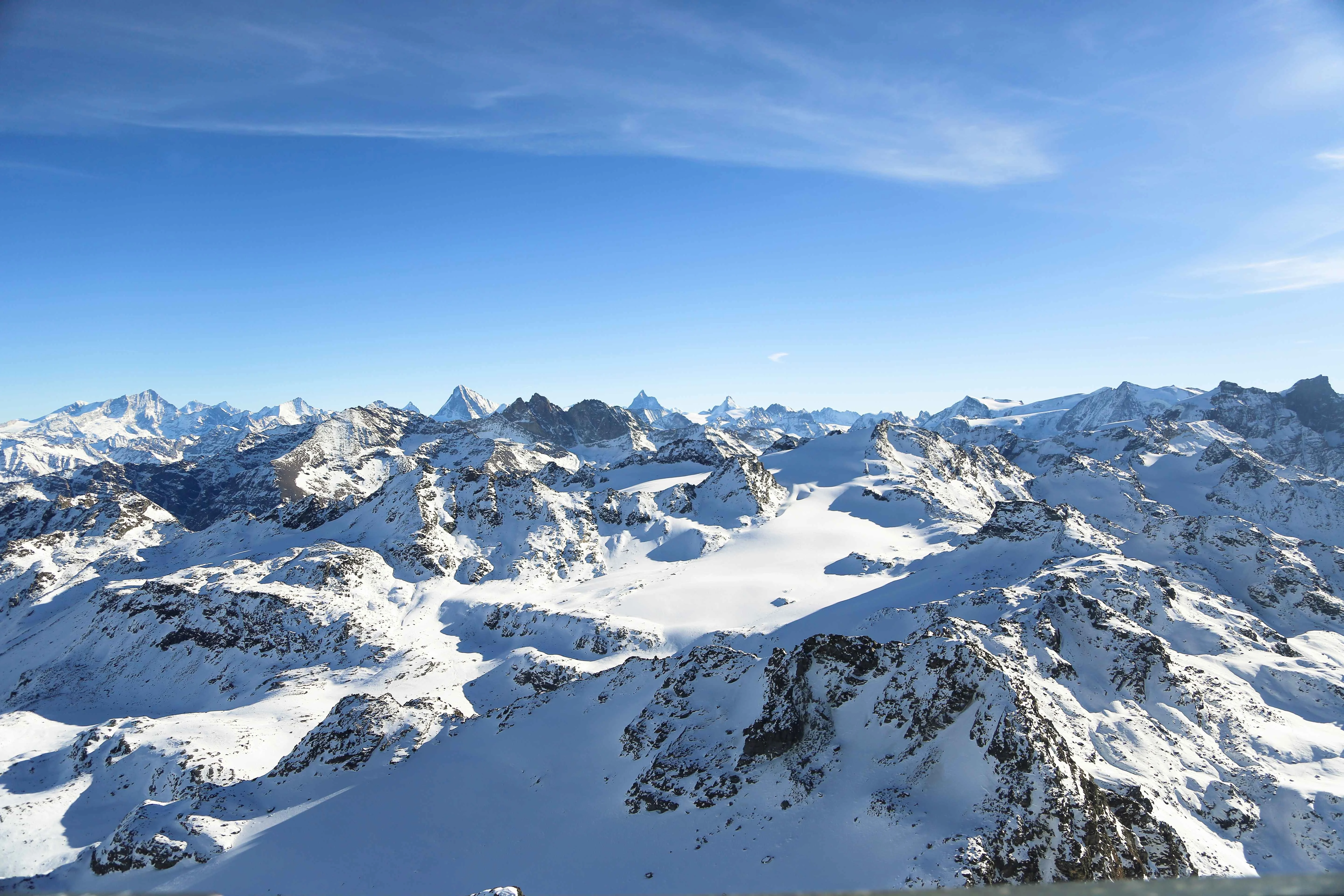 Snow-covered peaks and valleys of the Swiss Alps under a clear blue sky, featuring rugged terrain and distant mountain ranges.
