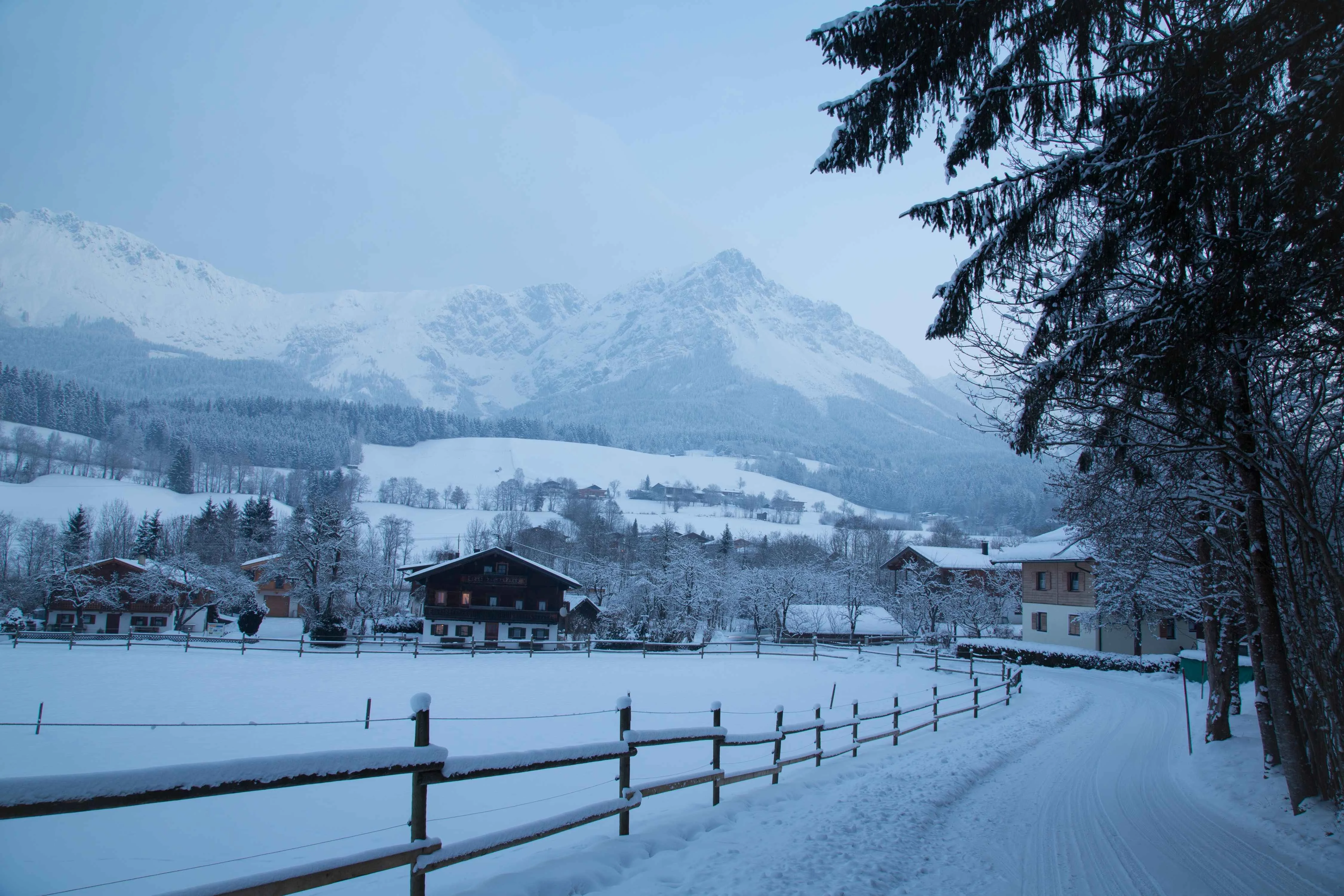 A serene winter scene in Scheffau, Austria, with snow-covered chalets, a winding road, and towering alpine peaks in the background under a soft blue sky