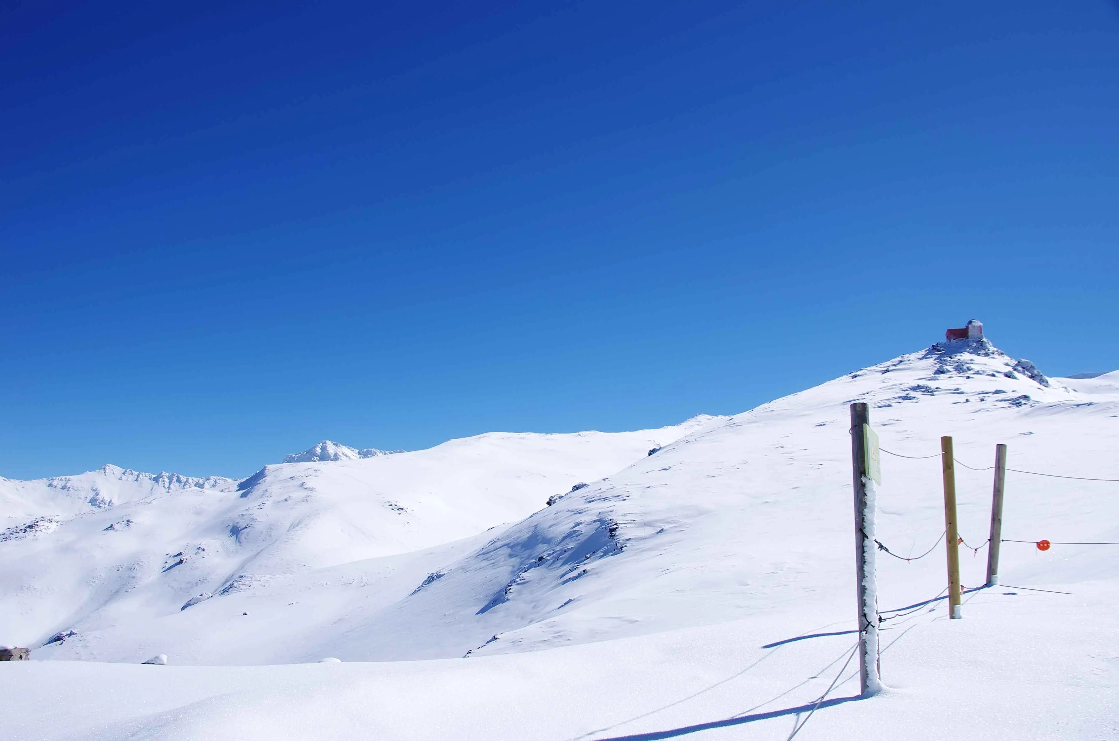 Snow-covered slopes of the Sierra Nevada mountains in Spain under a clear blue sky, with a small structure visible on a distant peak.
