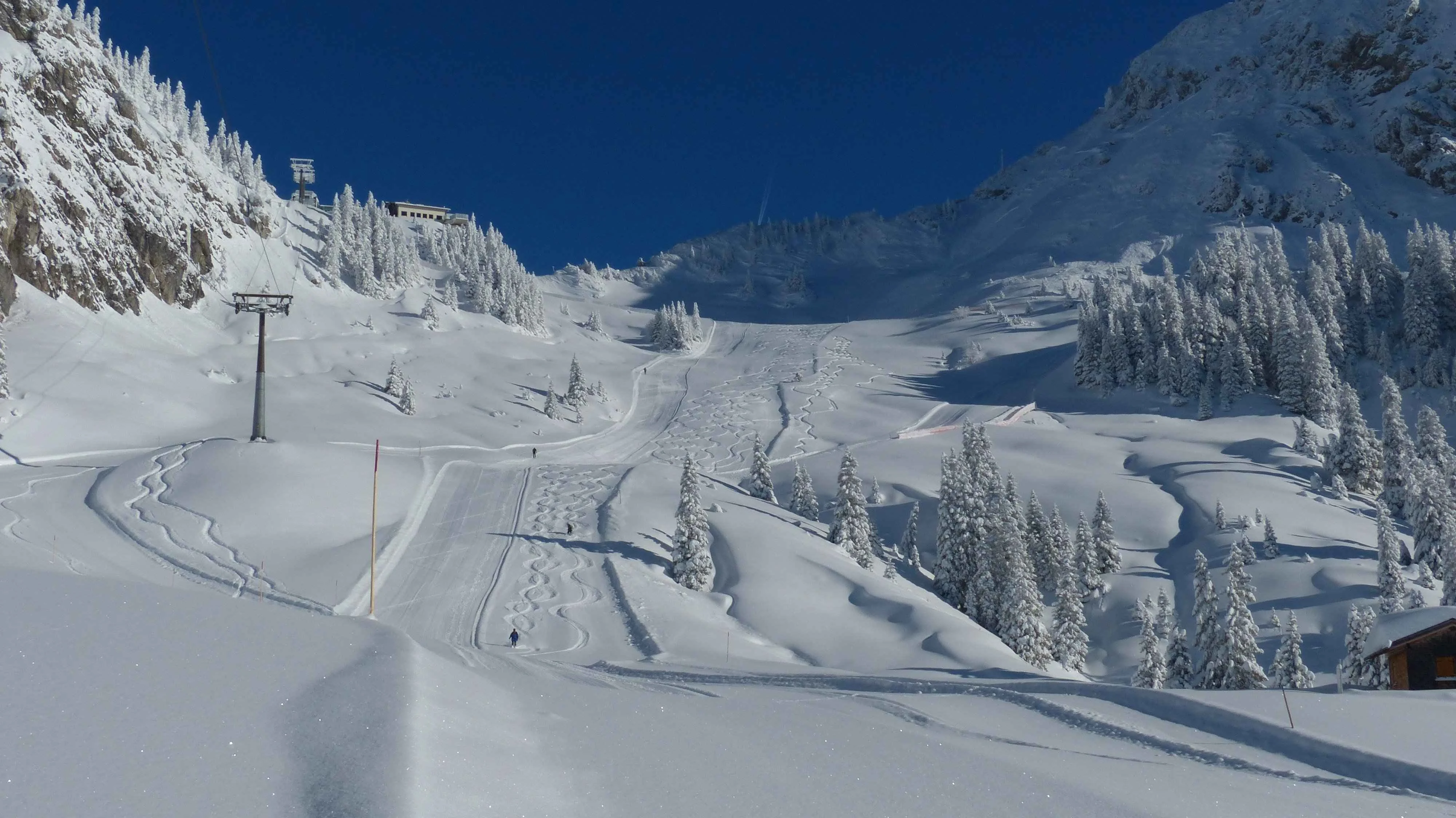 freshly groomed ski slope with ski tracks winding through powder snow, surrounded by snow-covered trees and a chairlift under a deep blue sky.
