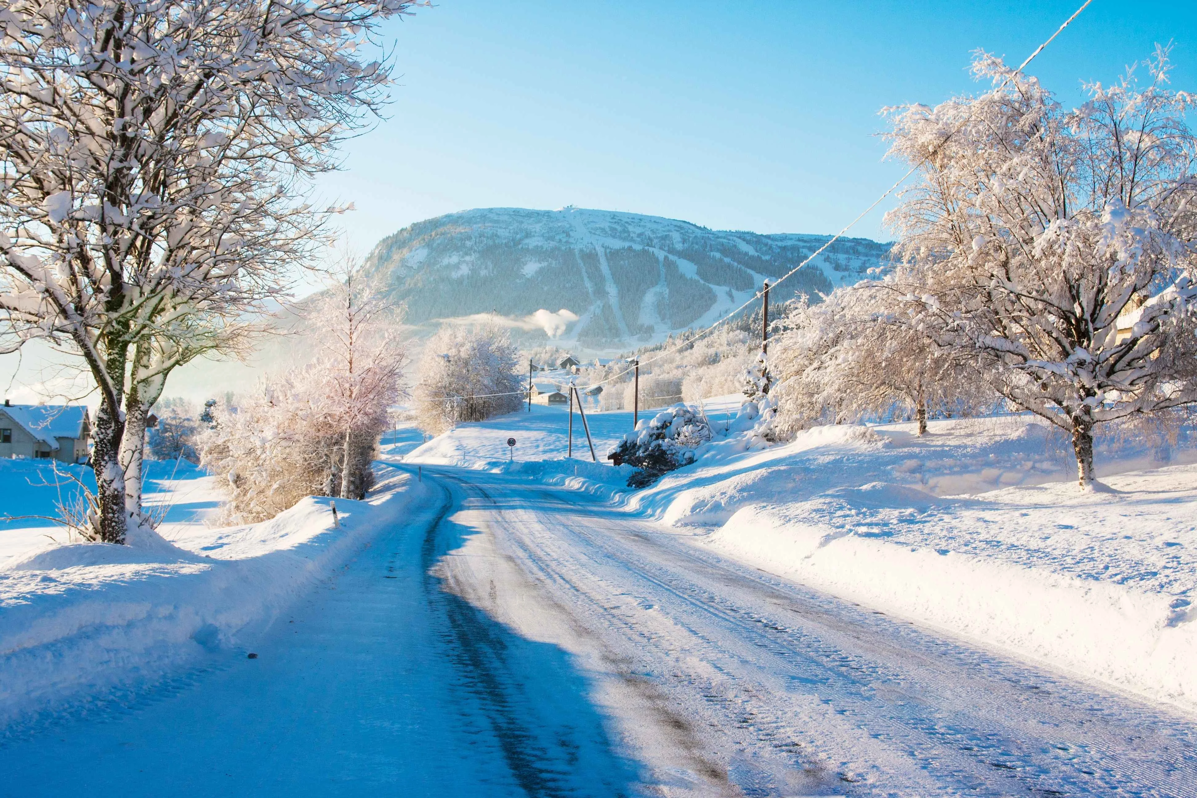 A snow-covered country road winds through a wintry landscape in Voss, Norway, with frost-coated trees on both sides and ski slopes visible on the forested mountain in the distance under a clear blue sky