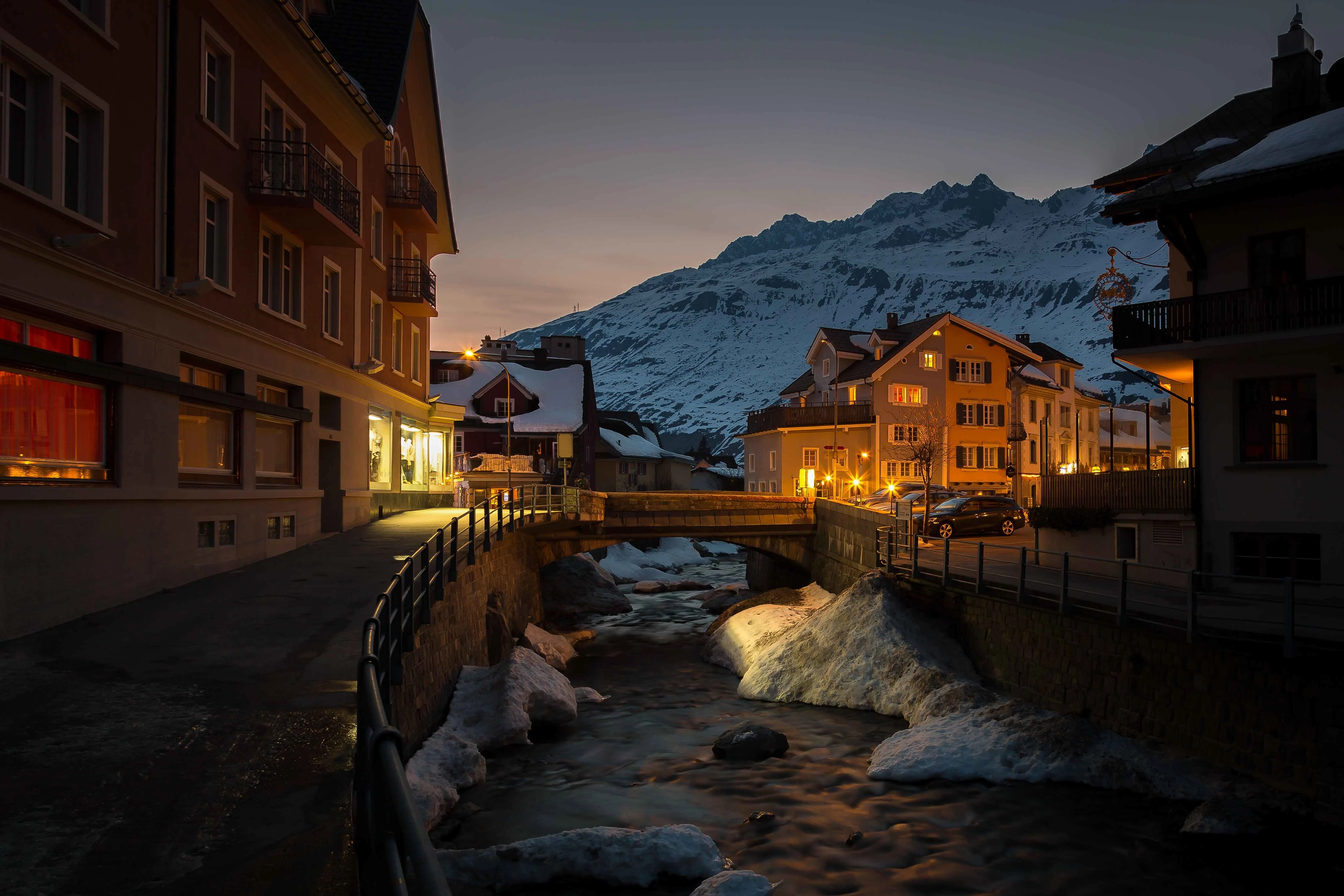 A charming winter evening in Andermatt, Switzerland, with warmly lit buildings lining a narrow stream, snow-covered mountains rising in the background, and soft twilight casting a peaceful glow over the alpine village.