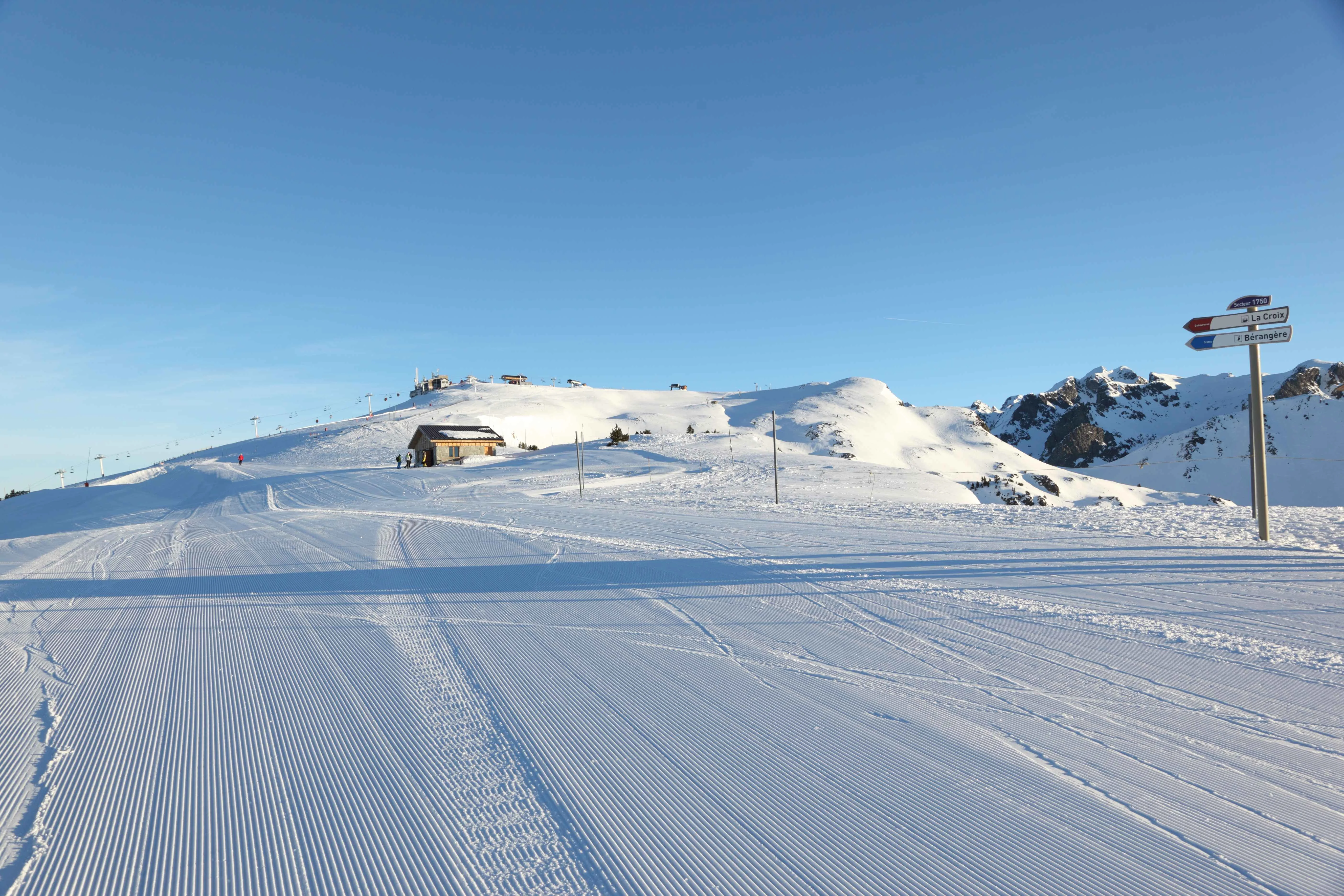 Freshly groomed ski slopes under a clear blue sky in Chamrousse, France, with mountain huts, ski lift towers, and piste signposts visible in the distance.