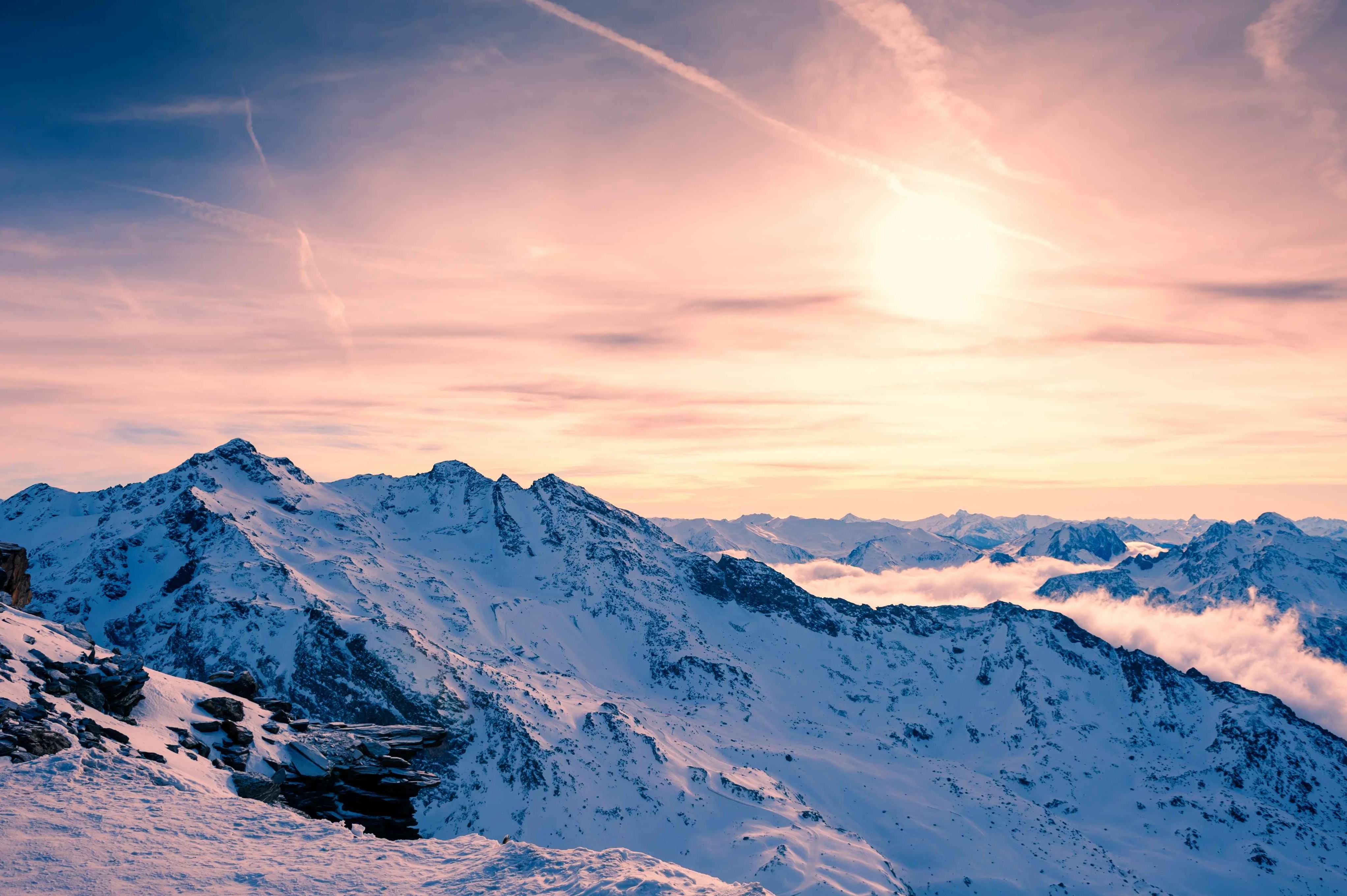 Panoramic view of the Three Valleys ski area at sunset, with snow-covered alpine peaks, dramatic ridgelines, and a glowing sky above a sea of clouds.