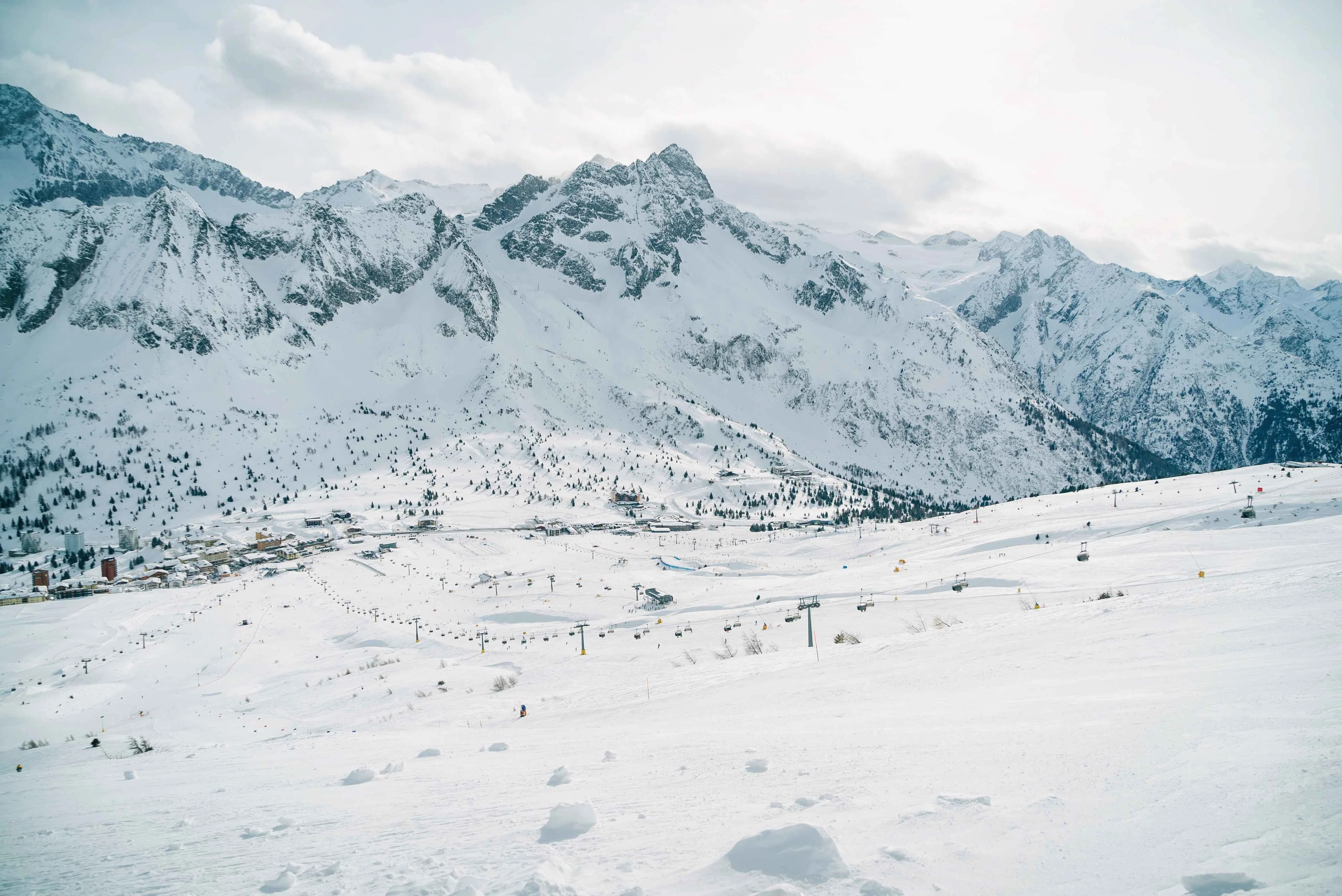 Panoramic view of Passo Tonale in Val di Sole, with wide snowy slopes, multiple chairlifts, and a cluster of ski resort buildings at the base, framed by dramatic Alpine peaks in the background.