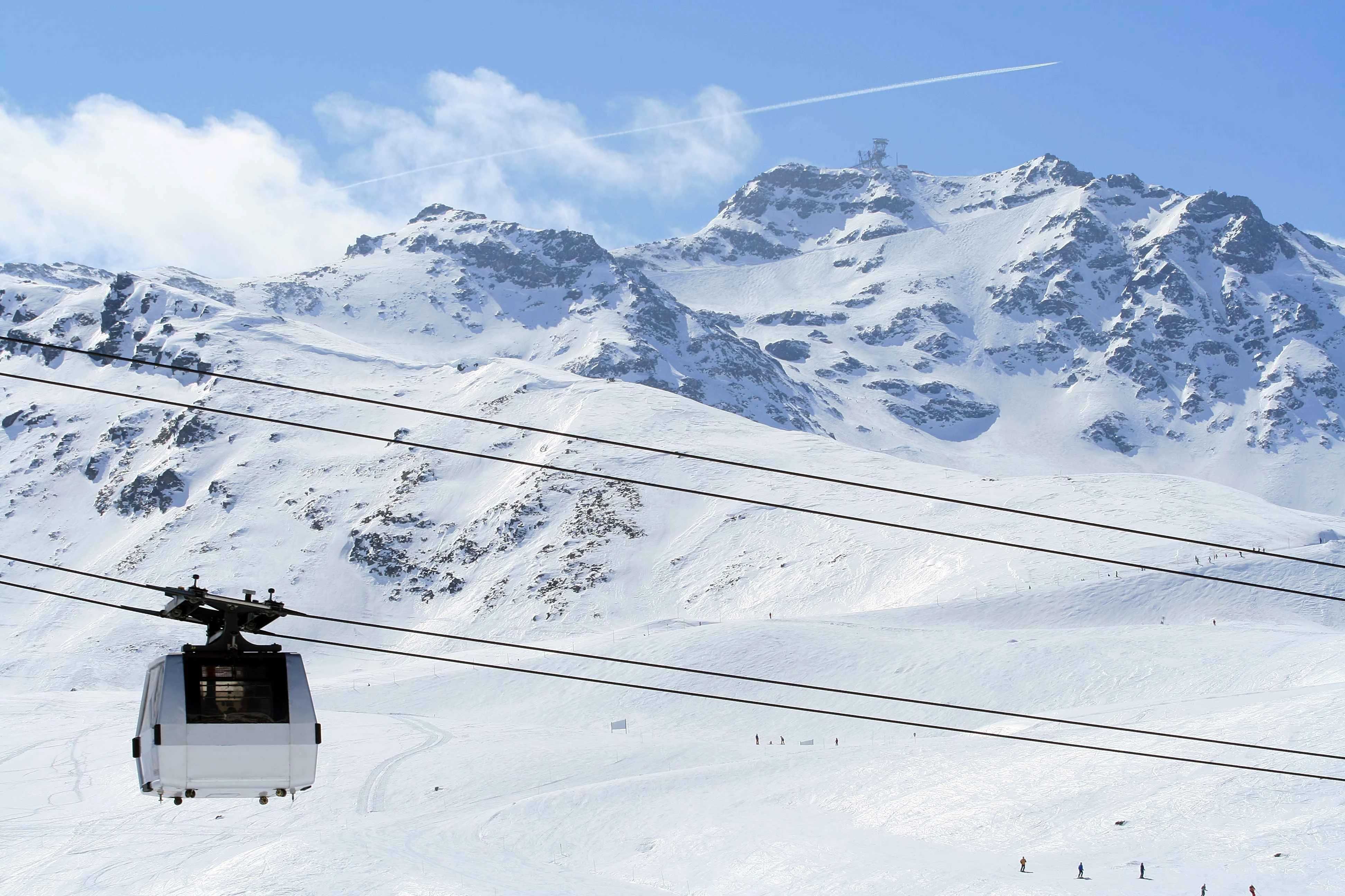 Close-up of a gondola lift gliding across snowy slopes with skiers below and rugged, sunlit mountain peaks rising in the background under a clear blue sky.