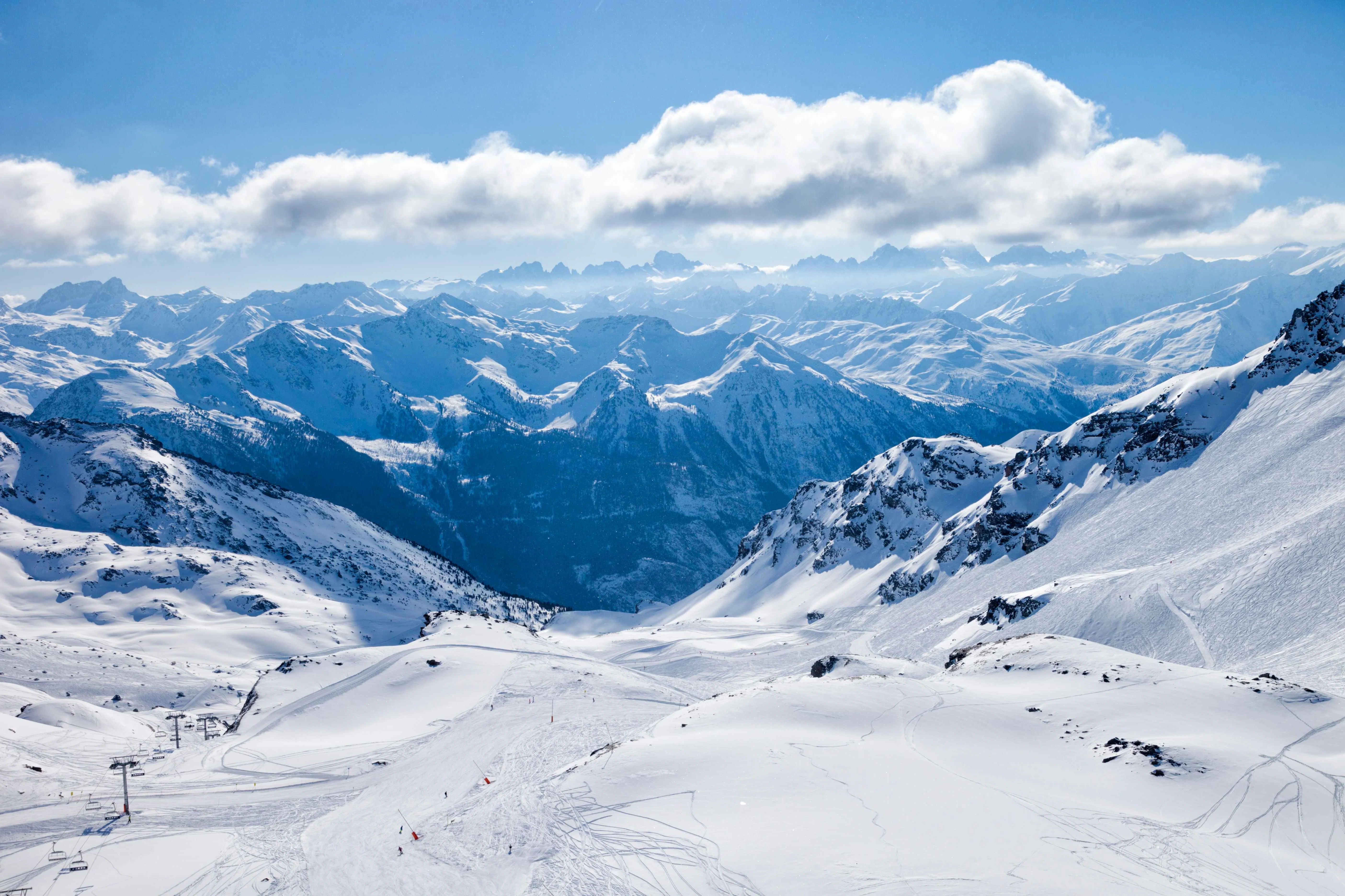 Panoramic view of the French Alps with vast snowy slopes, scattered ski lifts, and distant jagged peaks under a brilliant blue sky and drifting clouds.