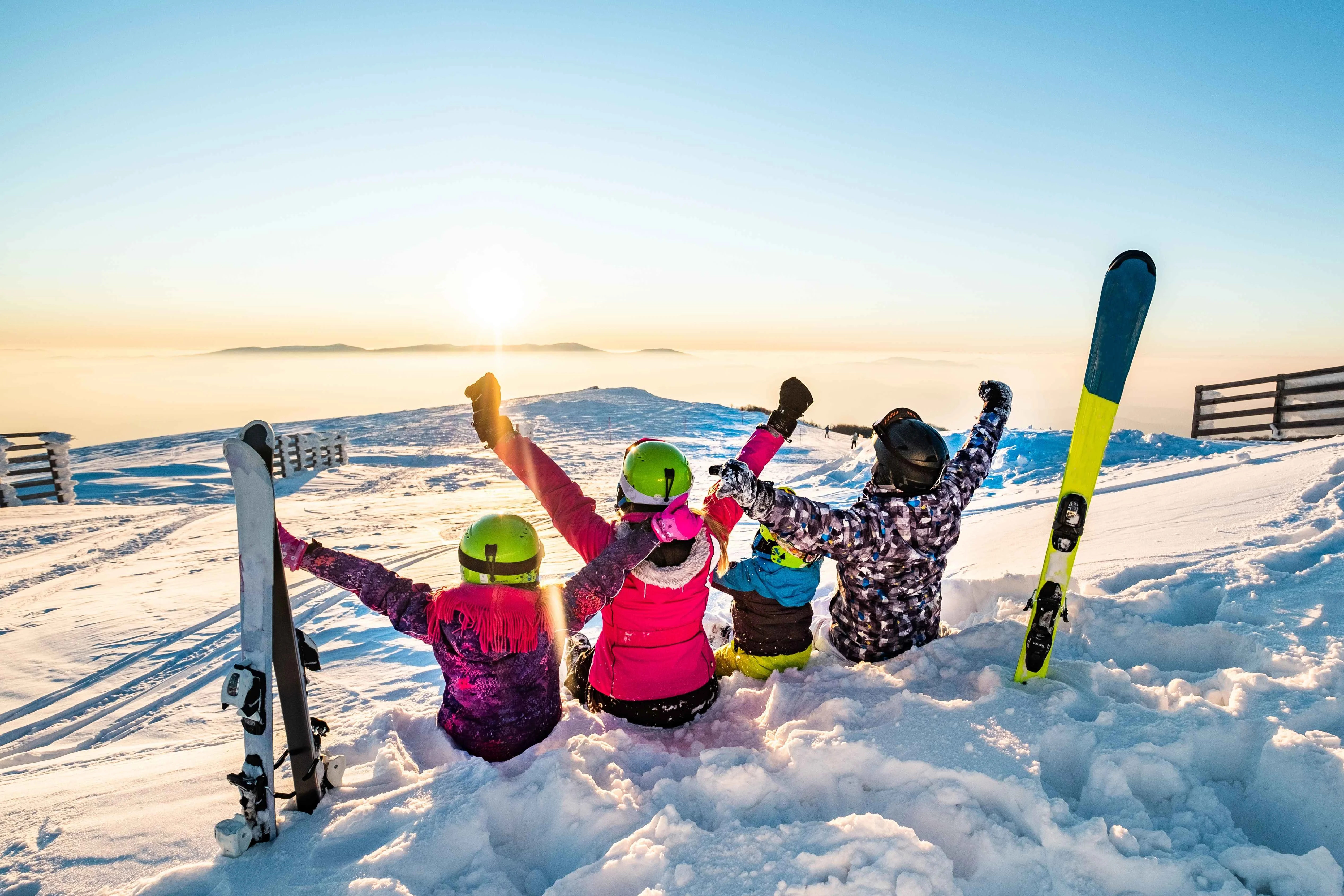 Happy family of four sitting in the snow with raised arms, celebrating a stunning sunset over snow-covered mountains during their ski holiday.