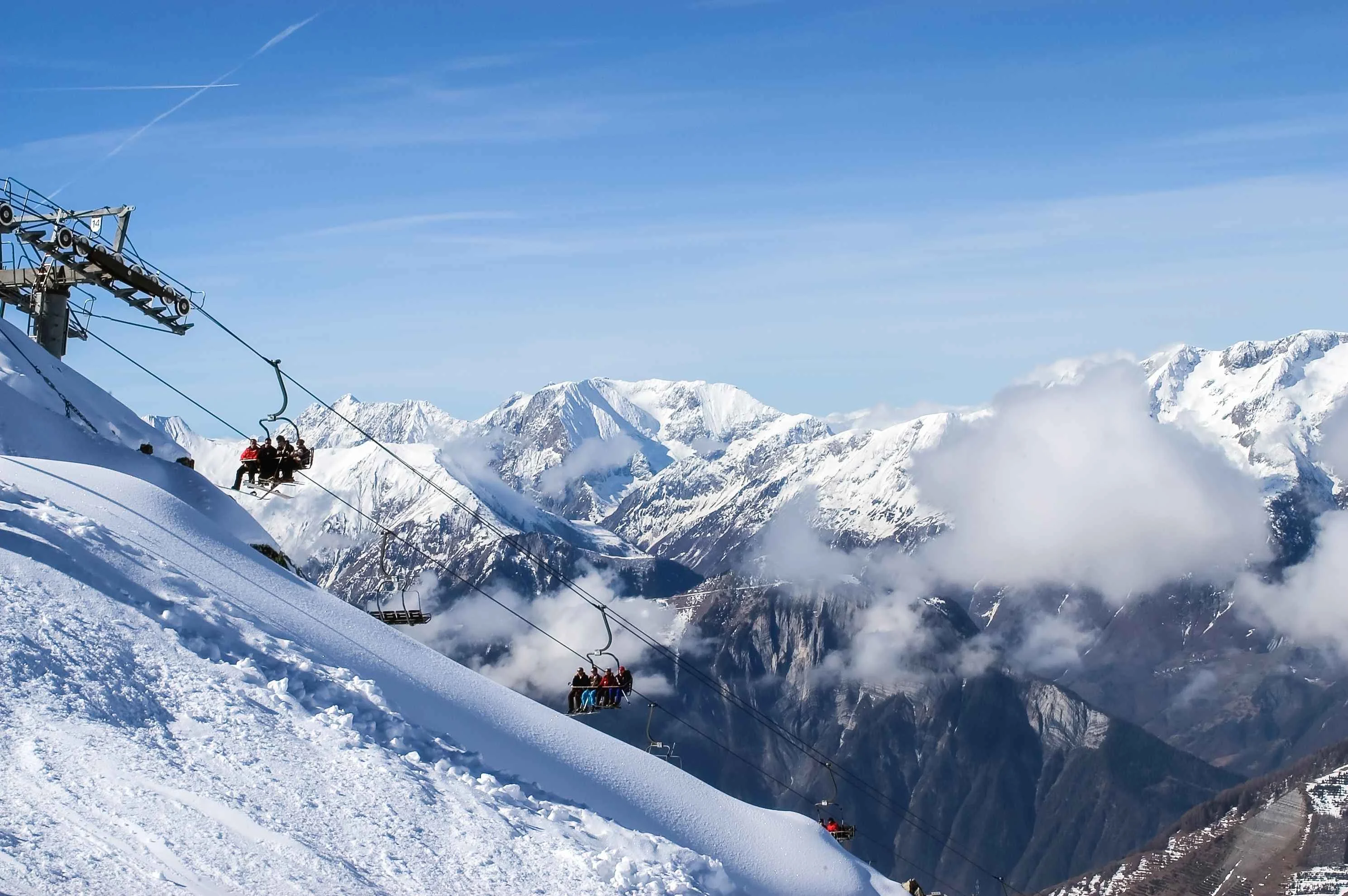 Chairlift ascending above Alpe d'Huez’s snowy ridgeline, with skiers riding up toward the slopes and panoramic views of the French Alps stretching into the distance.