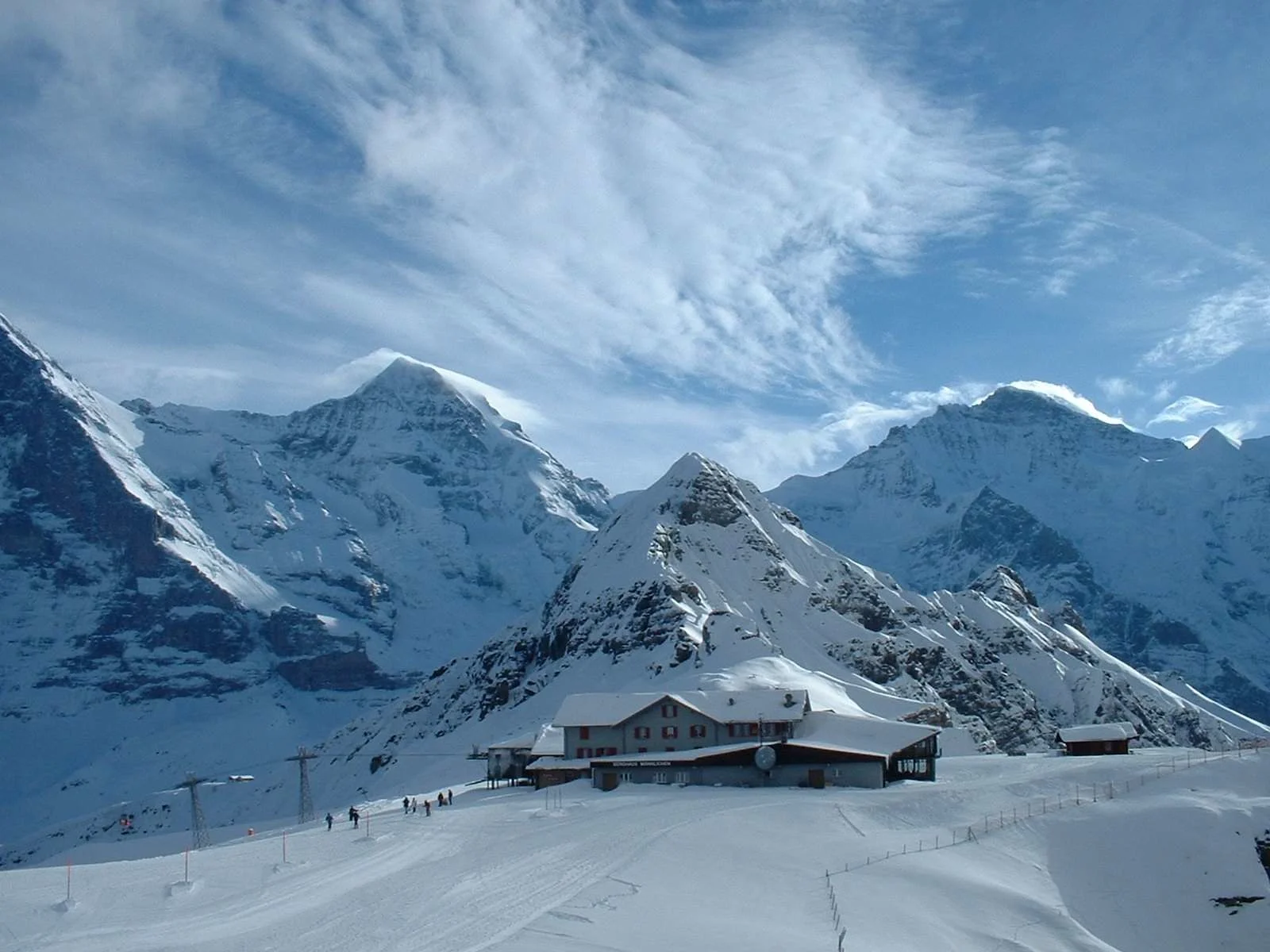 Snow covered ski lift nestled in the Swiss Alps with skiers on the slope below, framed by dramatic peaks and a sweeping alpine skyline under a crisp blue sky.