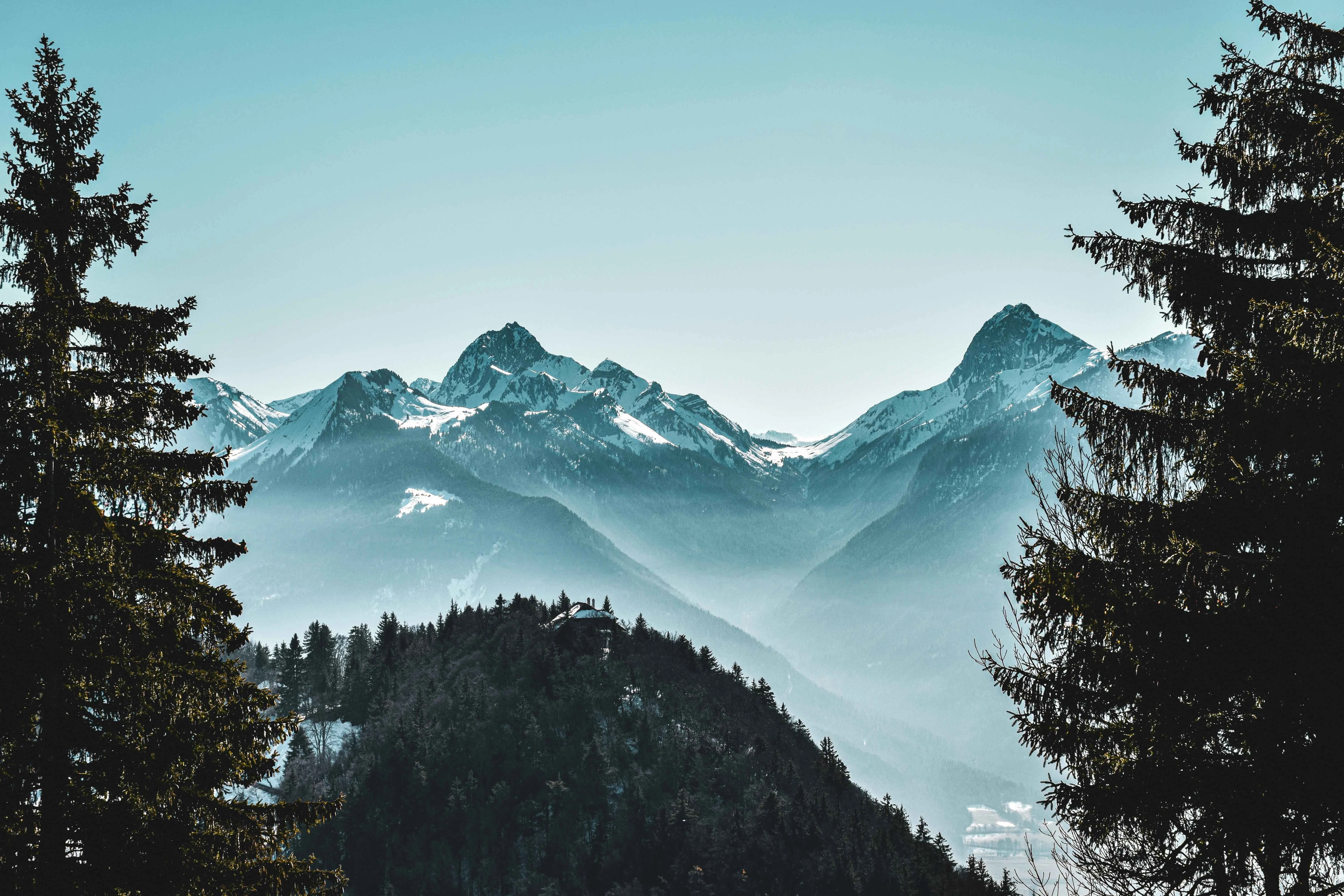 Snow-dusted alpine peaks surrounding Chamonix, framed by dark pine trees with morning mist layering the valleys for a dramatic and serene mountain view.