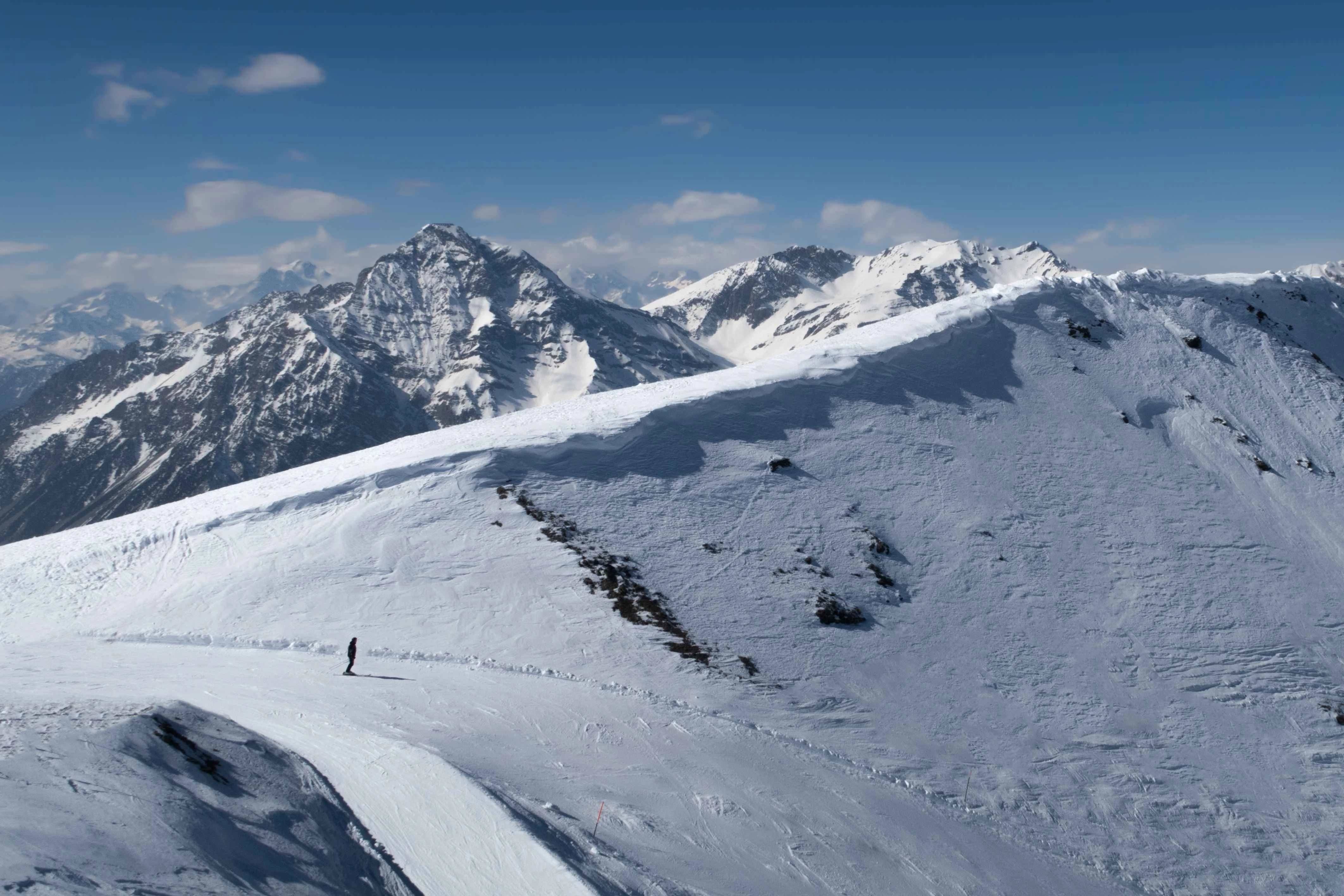 A lone skier carves across a quiet Sestriere ridgeline beneath the dramatic peaks of the Cottian Alps, where open slopes and rugged summits stretch endlessly beneath a crisp blue sky