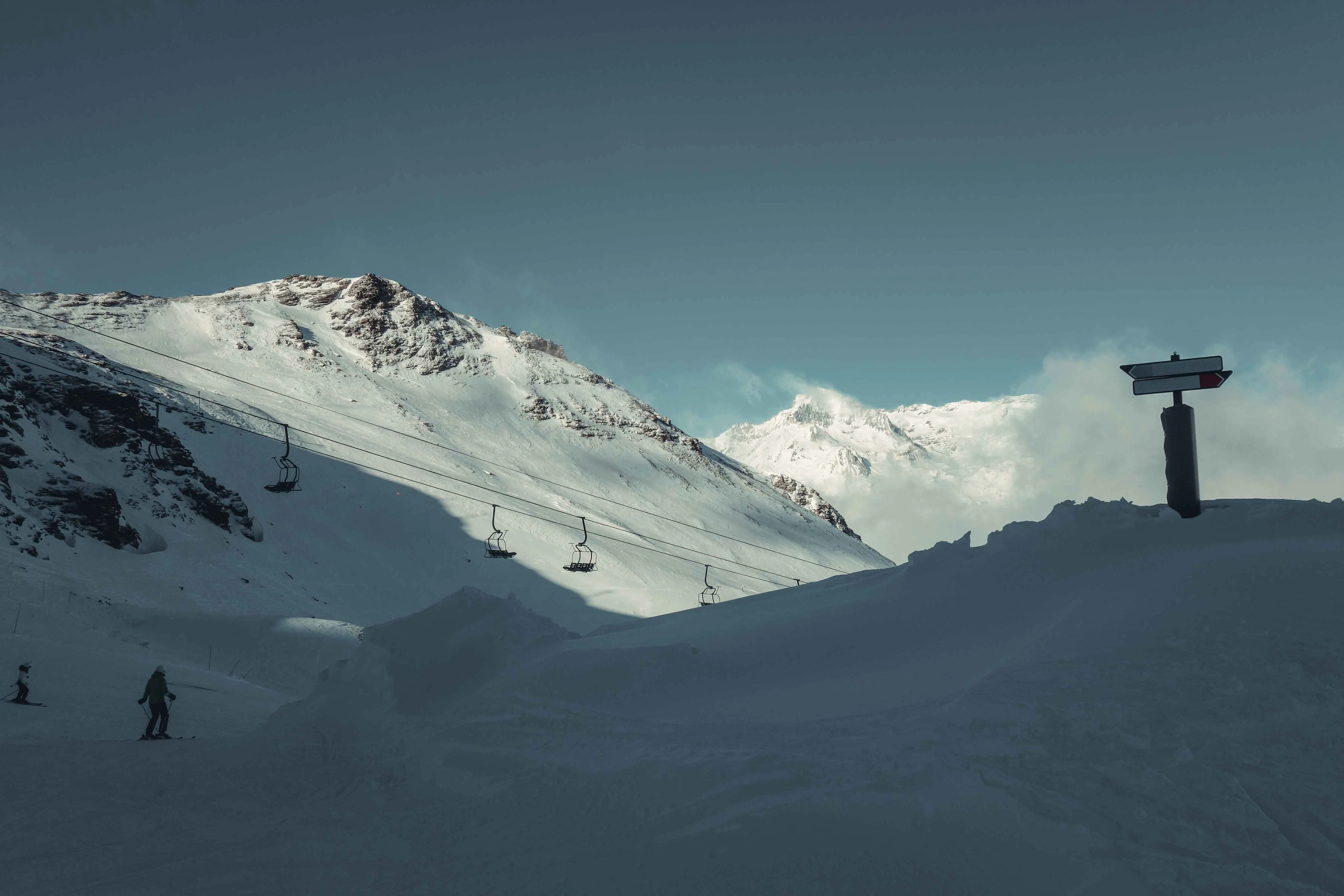 Skis point downhill beneath the morning shadow at Val Cenis, where empty chairlifts glide above freshly groomed pistes, framed by jagged alpine peaks glowing softly in the winter light